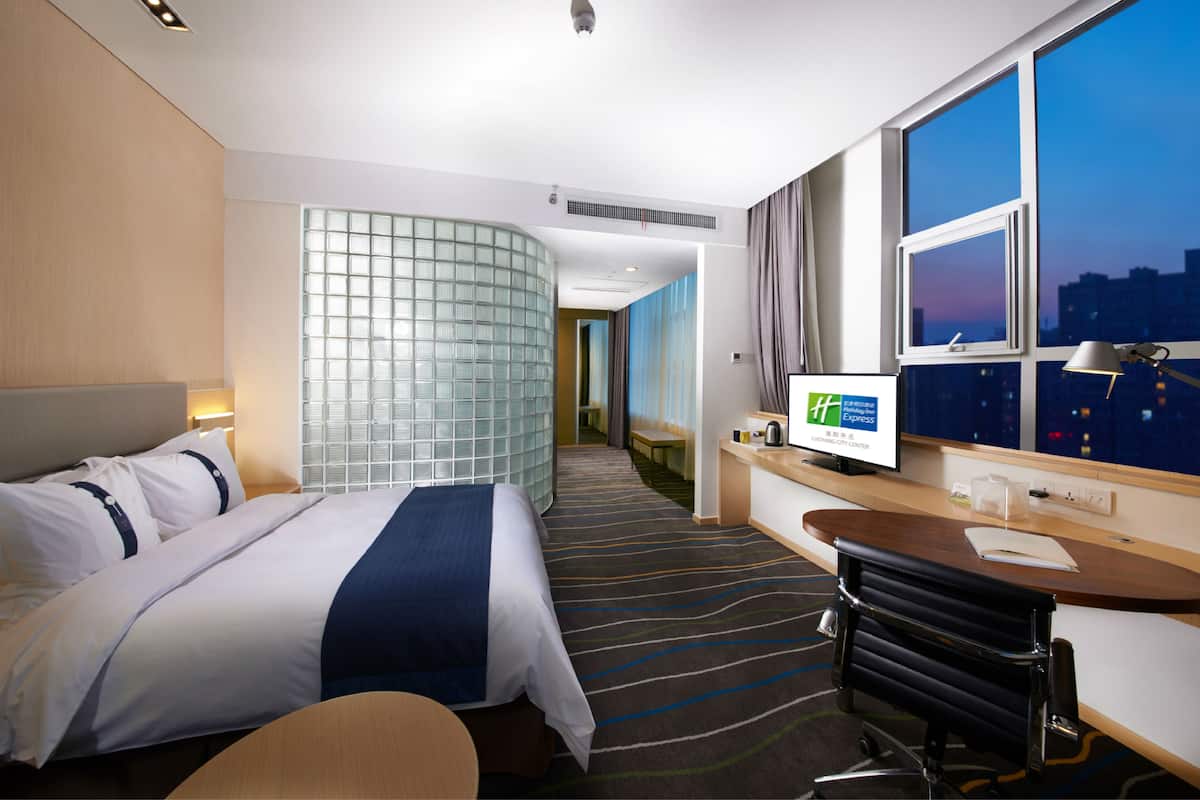 Ein Hotelzimmer mit Bett, Schreibtisch, Fernseher und großen Fenstern mit Blick auf die Stadt.