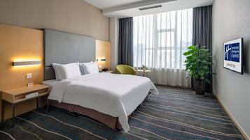 A hotel room with a large bed, bedside tables, a desk, a chair, a potted plant, and a window with curtains.