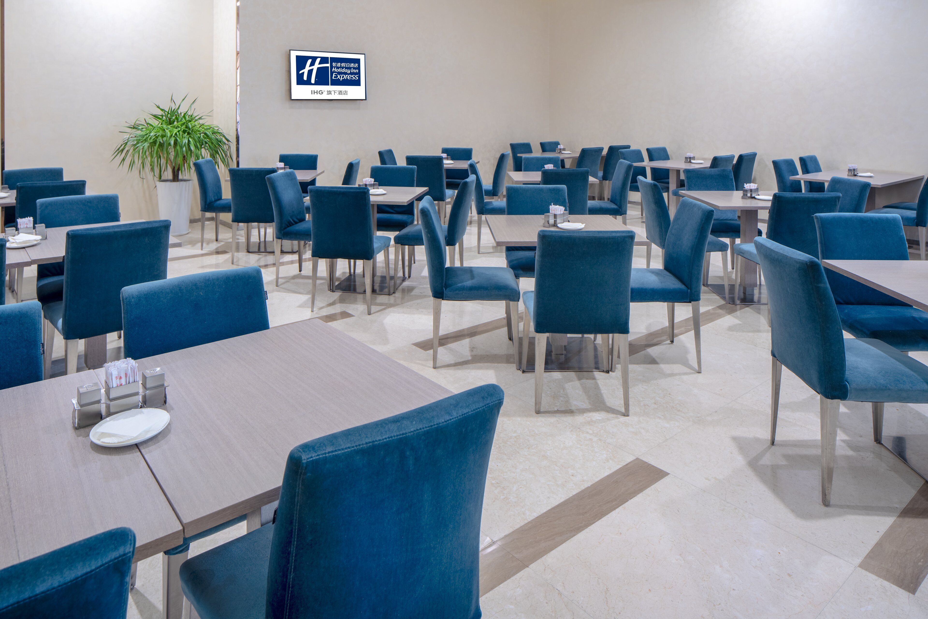 Un comedor de hotel con varias mesas y sillas, una planta en maceta y un cartel en la pared.