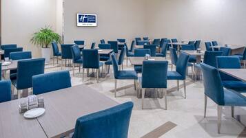 A hotel dining area with multiple tables and chairs, a potted plant, and a sign on the wall.