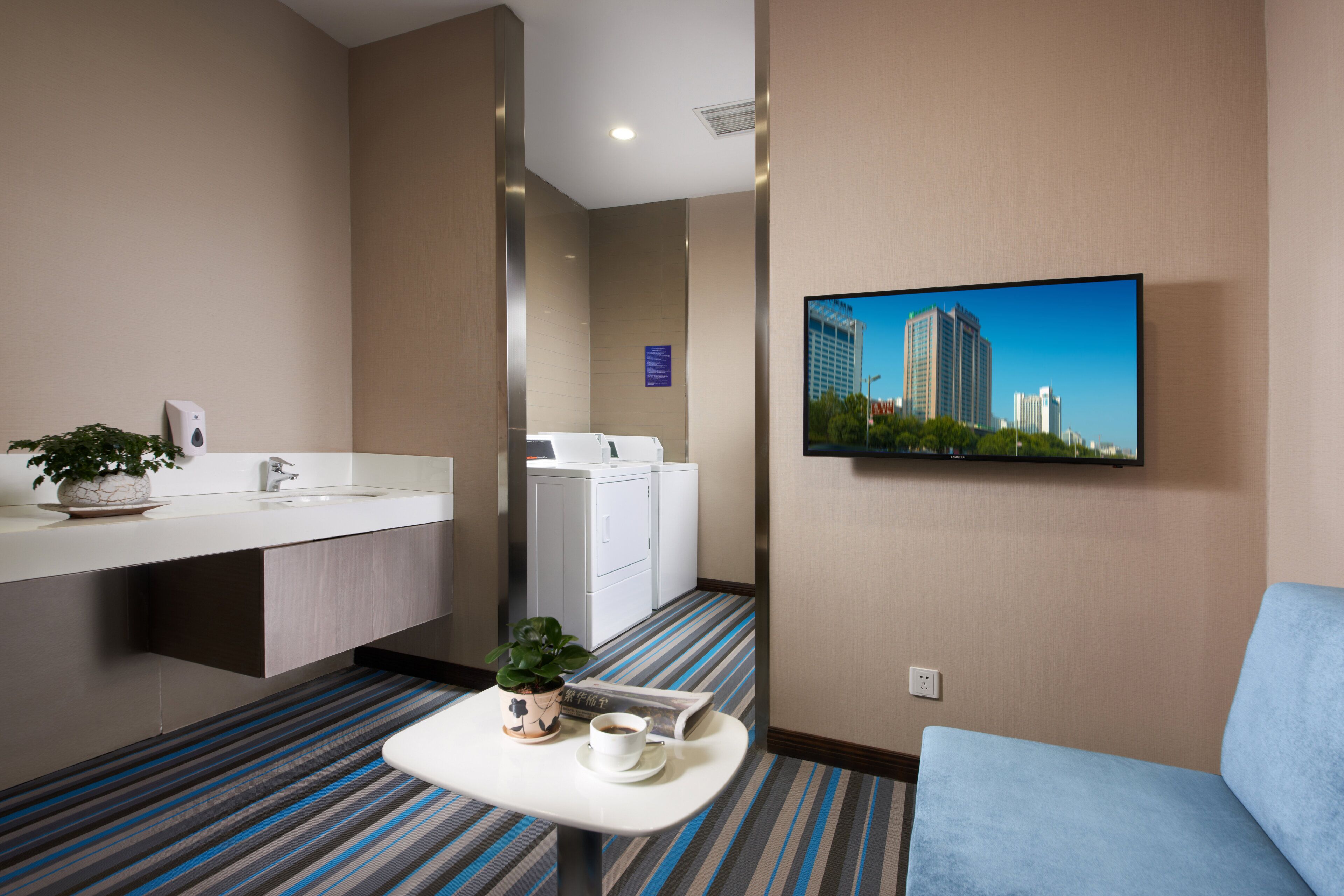 Habitación de hotel con un espejo grande, un lavamanos, una pizarra, un televisor de pantalla plana montado en la pared, una mesita con taza y platillo, y un sofá azul.