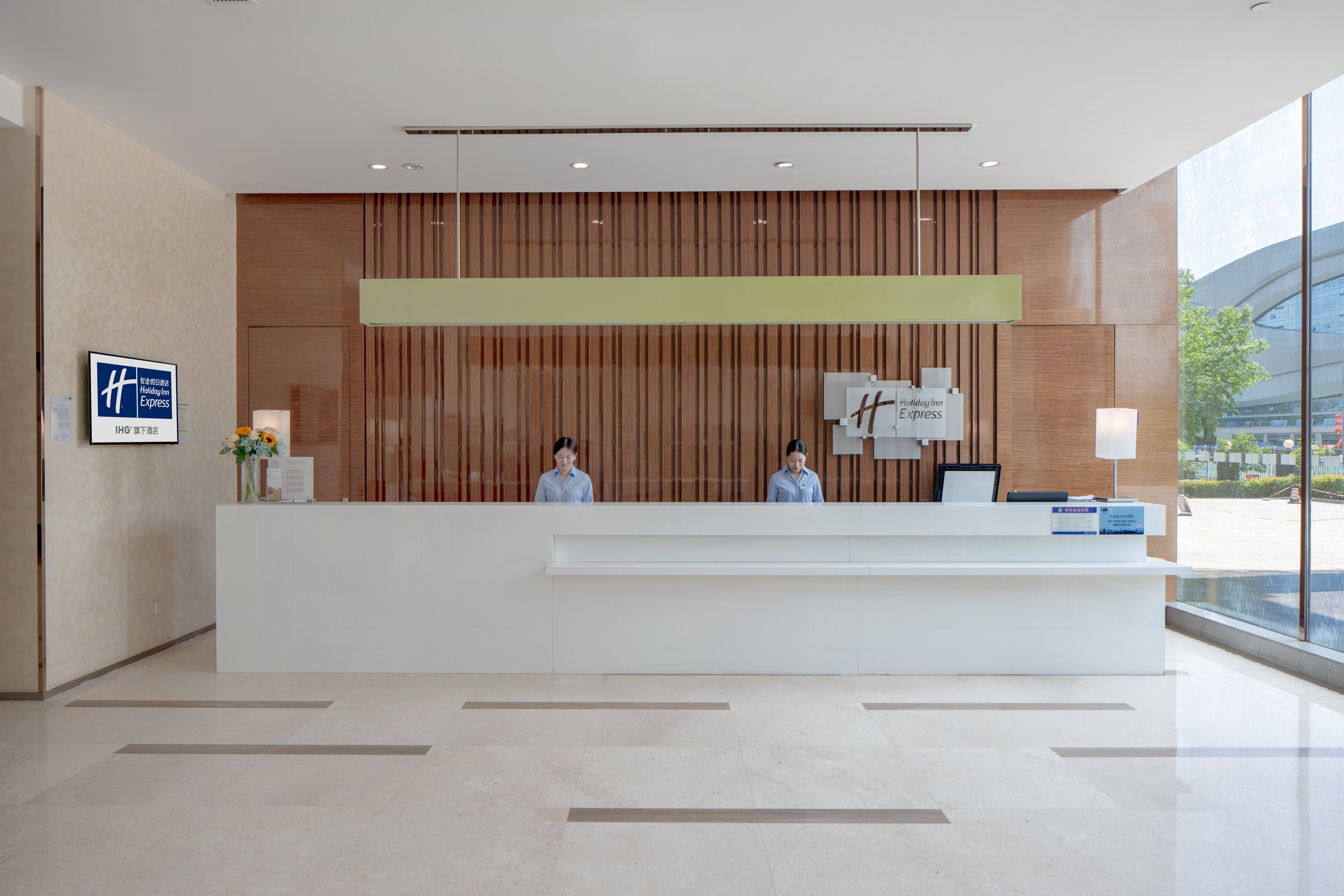 A hotel reception desk with two staff members, a wooden panel wall, and a large window offering a view of the city.