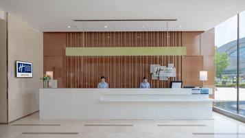 A hotel reception desk with two staff members, a wooden panel wall, and a large window offering a view of the city.