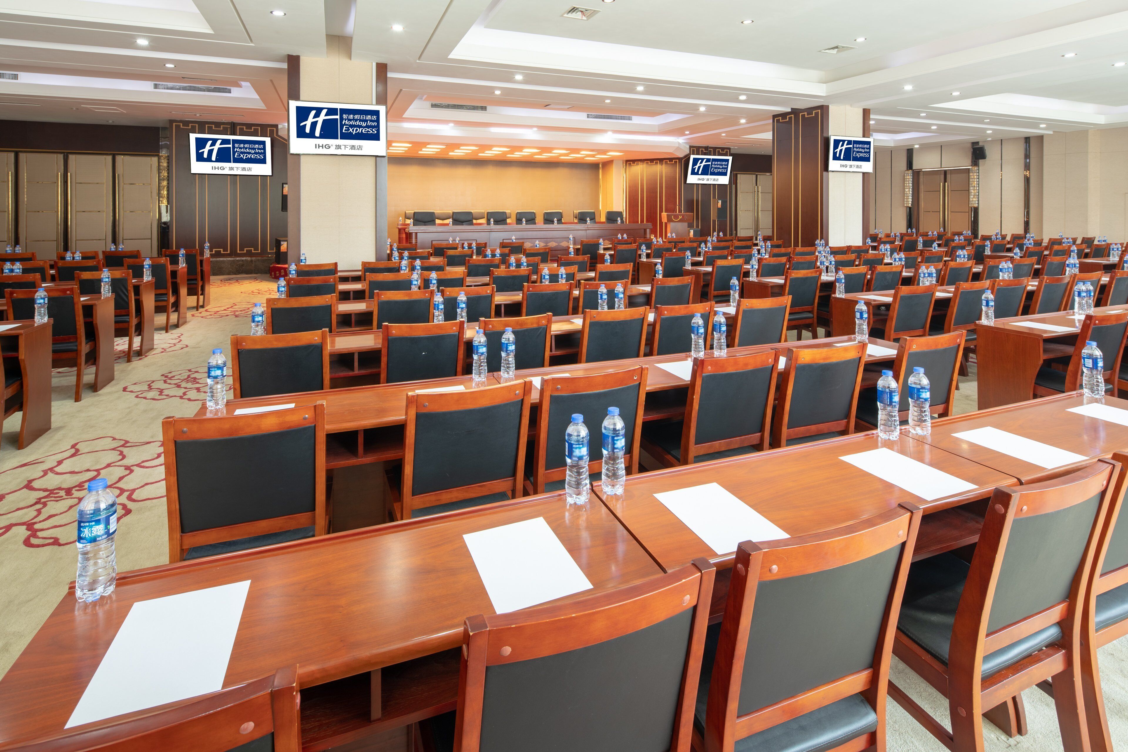 Un salón de conferencias con filas de mesas y sillas, equipado con botellas de agua, y señales que indican la ubicación de Holiday Inn Express.