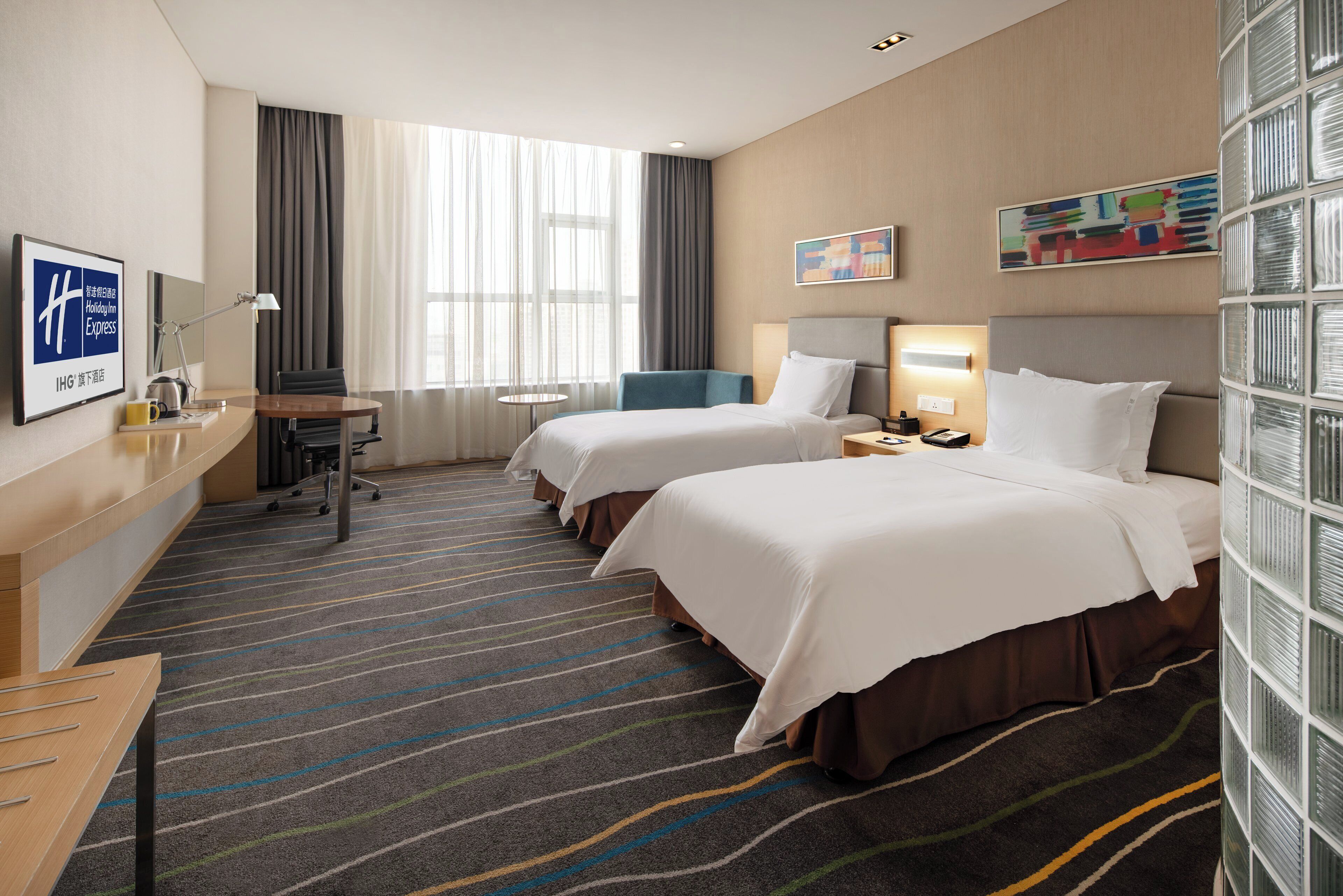 A hotel room with two beds, a desk, a chair, a small table, a lamp, and a window with curtains.
