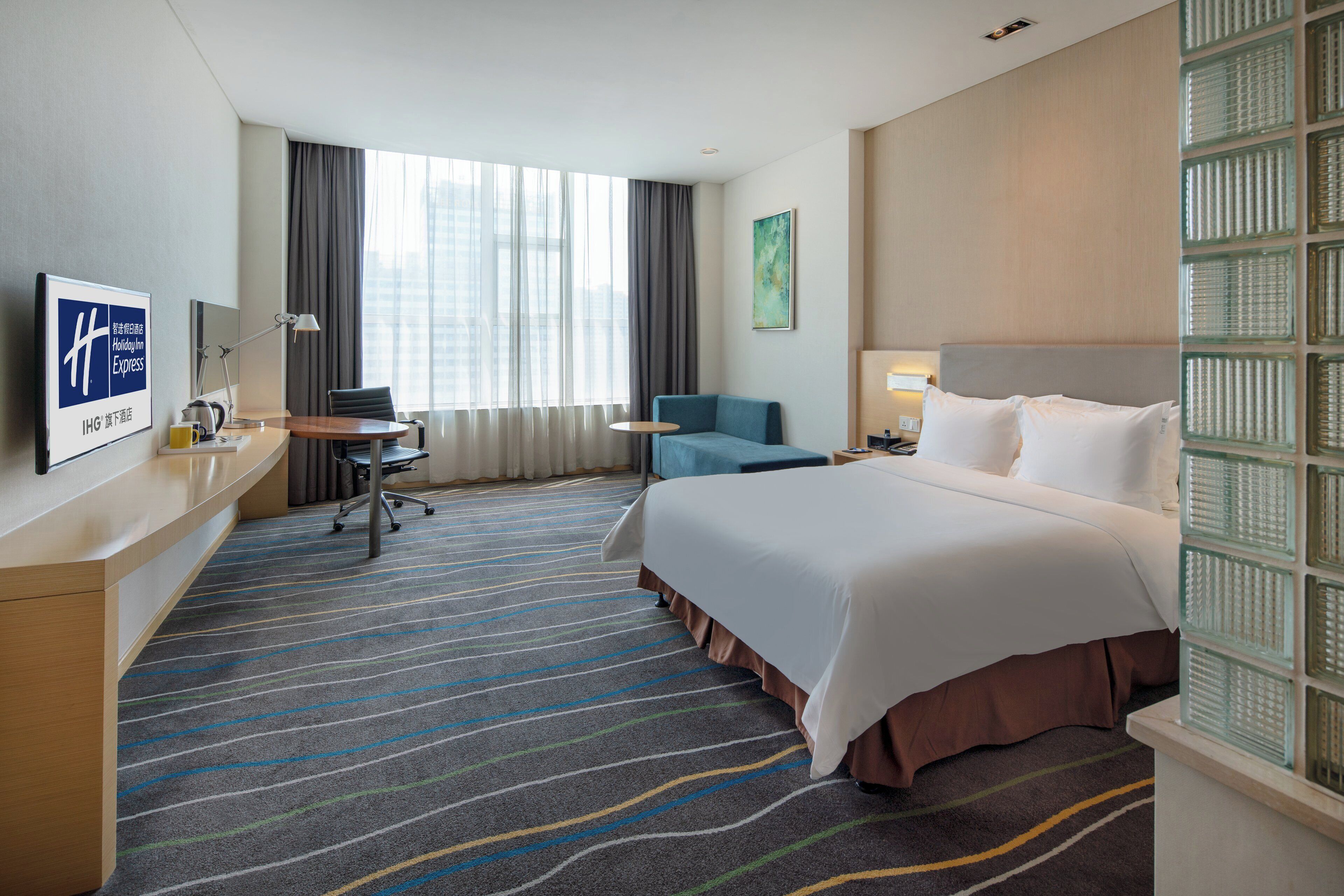 A hotel room with a bed, a desk, a chair, a window with curtains, and a wall-mounted artwork.