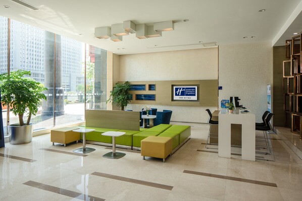 Zona de recepción de hotel con diseño moderno, que incluye un mostrador de recepción, un área de descanso con sofás y mesas, y un gran ventanal con vistas a la ciudad.