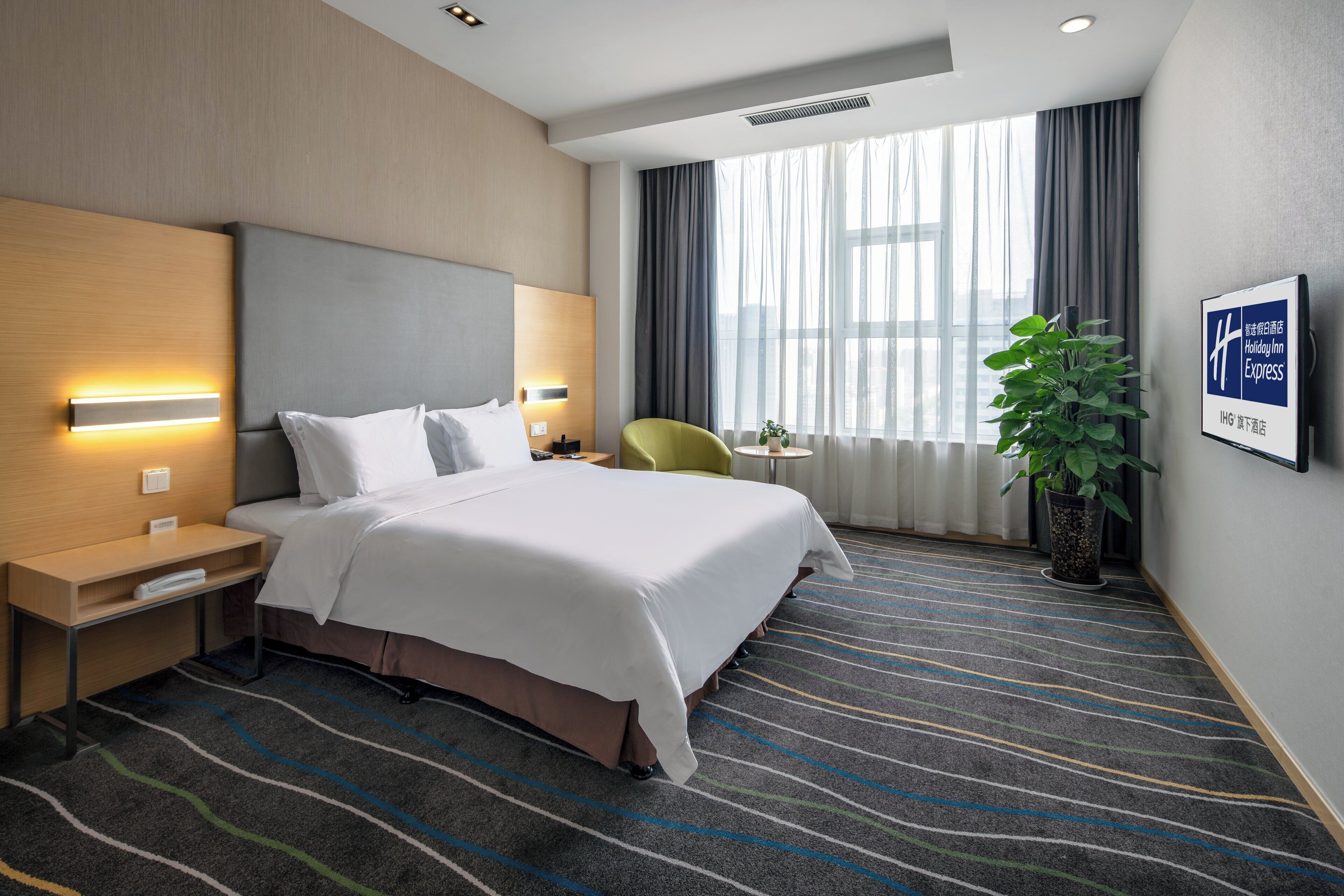 A hotel room with a bed, bedside tables, a chair, a potted plant, and a large window with curtains.
