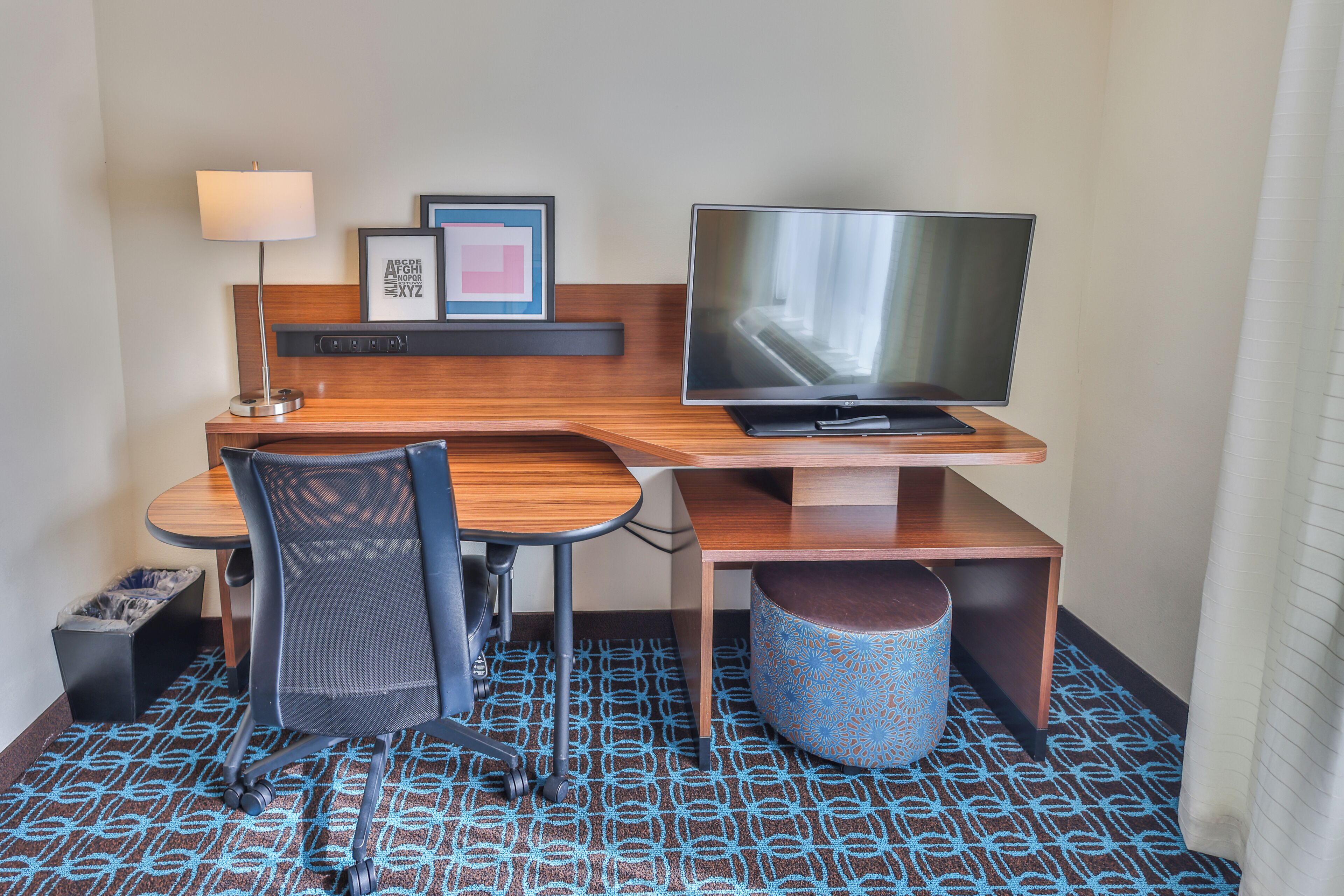 A hotel room with a wooden desk, a TV, a chair, a lamp, a trash bin, and a patterned carpet.