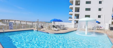 A swimming pool with lounge chairs and a fountain, surrounded by a fence and a building with balconies.