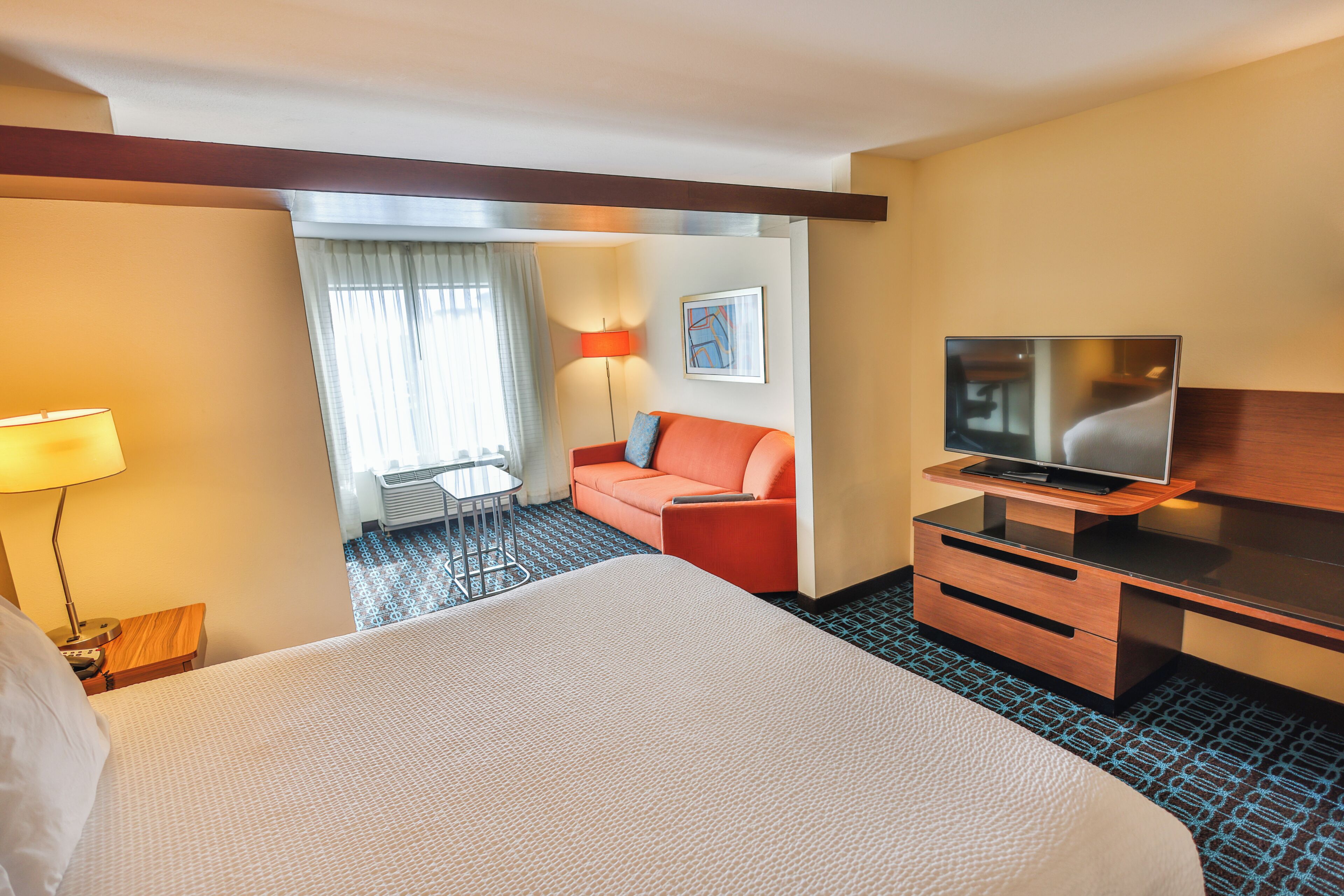 A hotel room with a large bed, a TV, a desk, a chair, a sofa, a lamp, and a window with curtains.