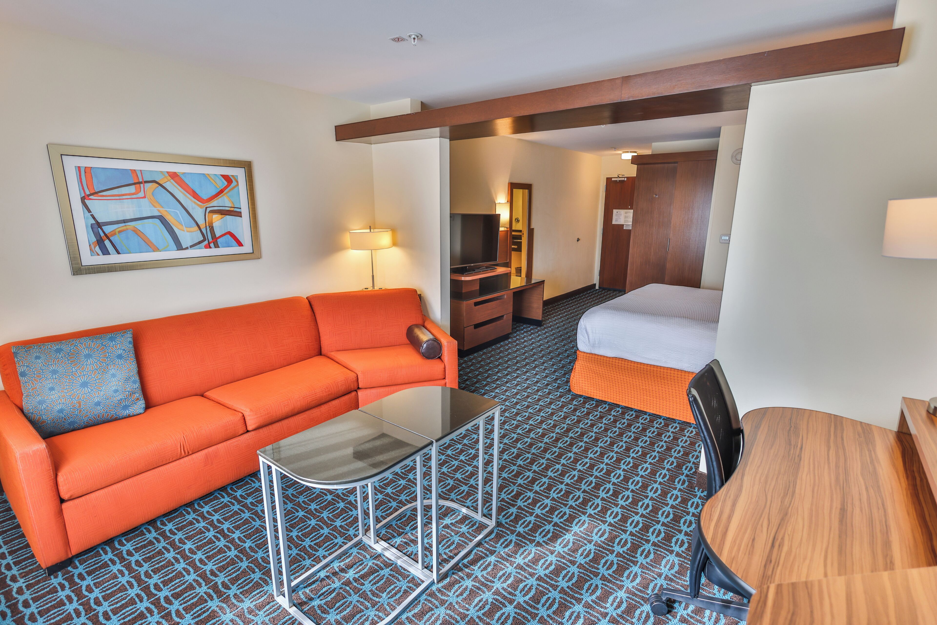 A hotel room with a sofa, a glass-top table, a bed, a wooden desk, and a TV.