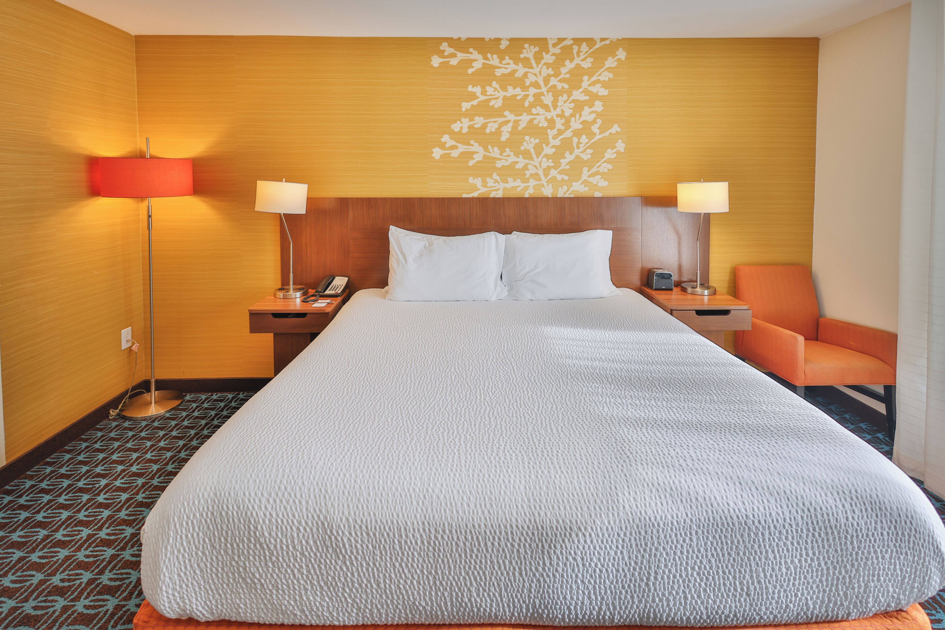 A hotel room with a large bed, two bedside tables, a nightstand, a lamp, and an orange chair.