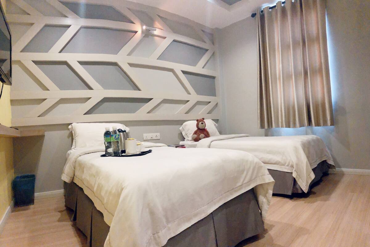 A hotel room with two beds, a decorative ceiling, a window with curtains, and a small trash bin.