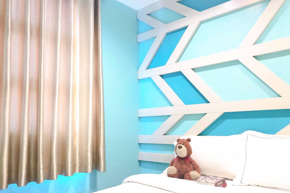 A bedroom with a bed, a teddy bear, a window with curtains, and a geometric wall design.