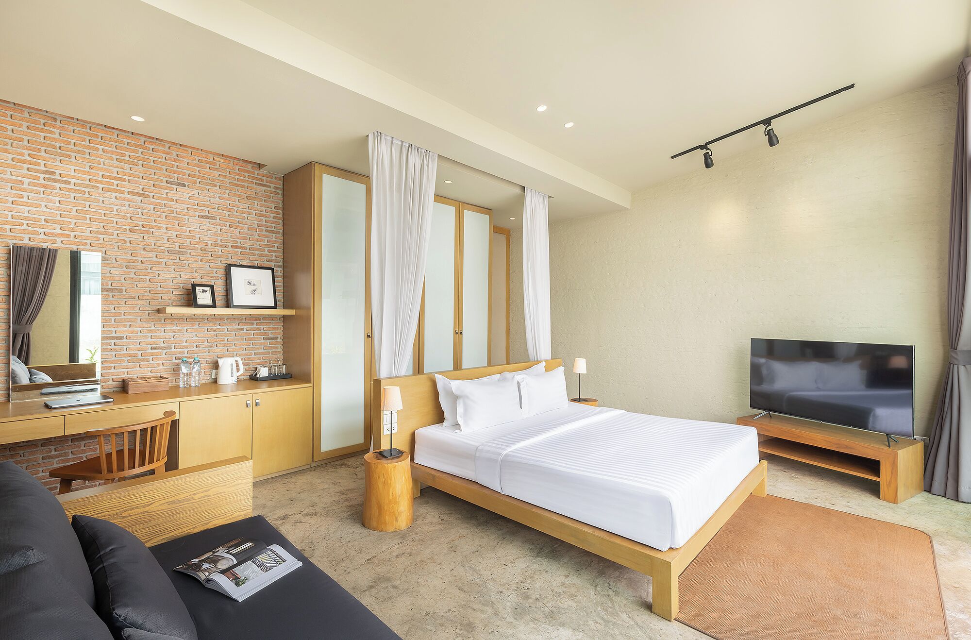 A modern hotel room with a brick accent wall, a large bed with white bedding, a TV, a desk with a computer, and a small seating area.
