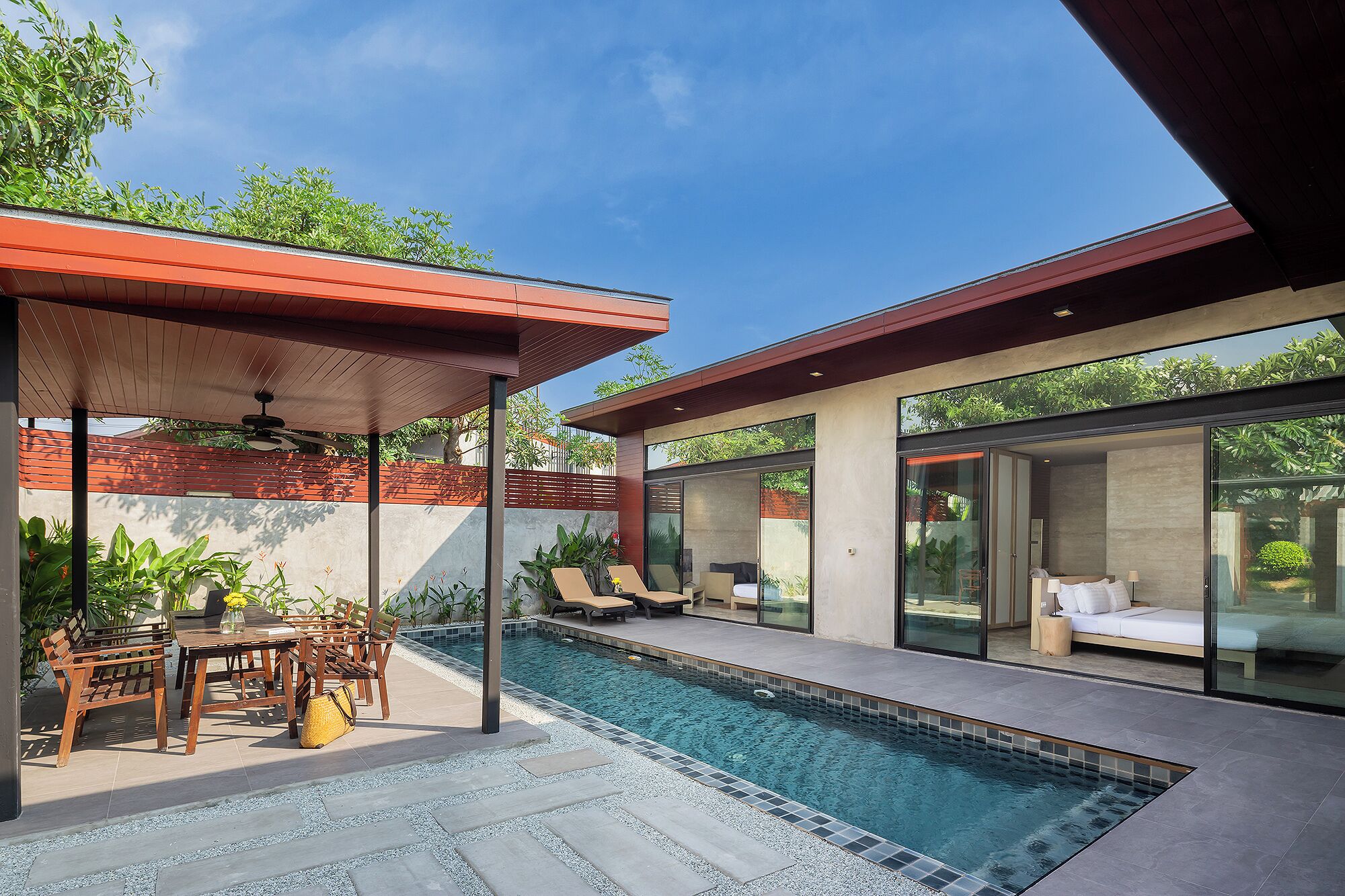 A modern outdoor pool area with a covered seating area, a glass-enclosed bedroom, and a view of the surrounding greenery.