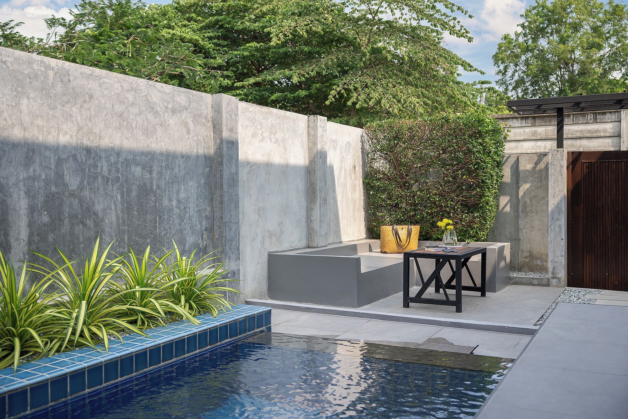 A modern outdoor pool area with a concrete wall, a small seating area with a table and chairs, and a potted plant.