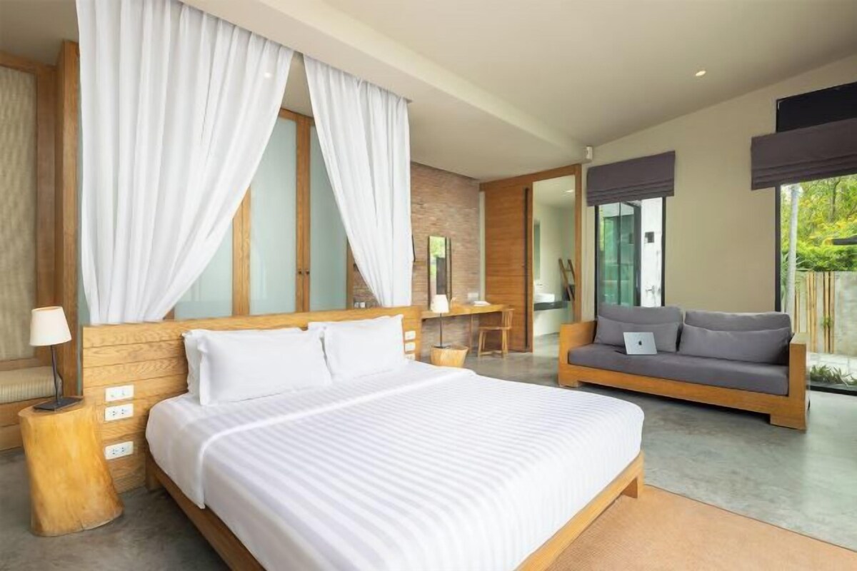 A modern hotel room with a large bed, a sofa, a desk, and a view of the outdoors.