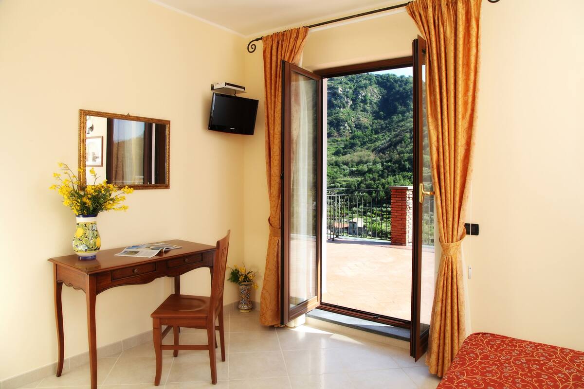 Quarto com escrivaninha de madeira, cadeira e uma mesinha com um vaso de flores. Há uma TV fixada na parede e uma porta de vidro deslizante que dá para uma área externa com vista para vegetação.
