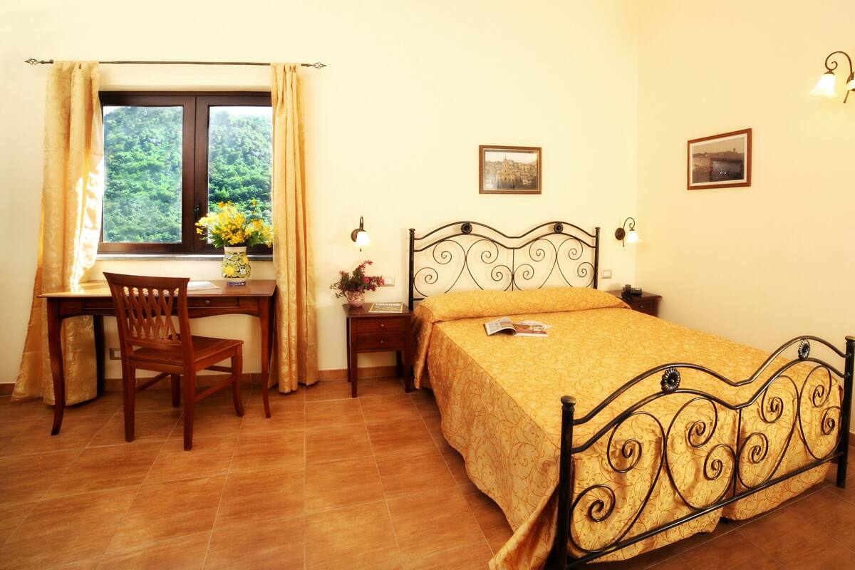 Quarto com cama, escrivaninha e cadeira. Há duas luminárias na parede e duas imagens emolduradas.