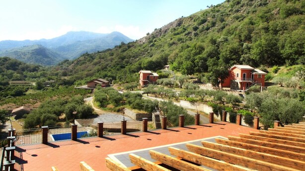 Propiedad en una ladera con un edificio de techo rojo, una terraza de madera y vistas a la vegetación circundante y montañas distantes.