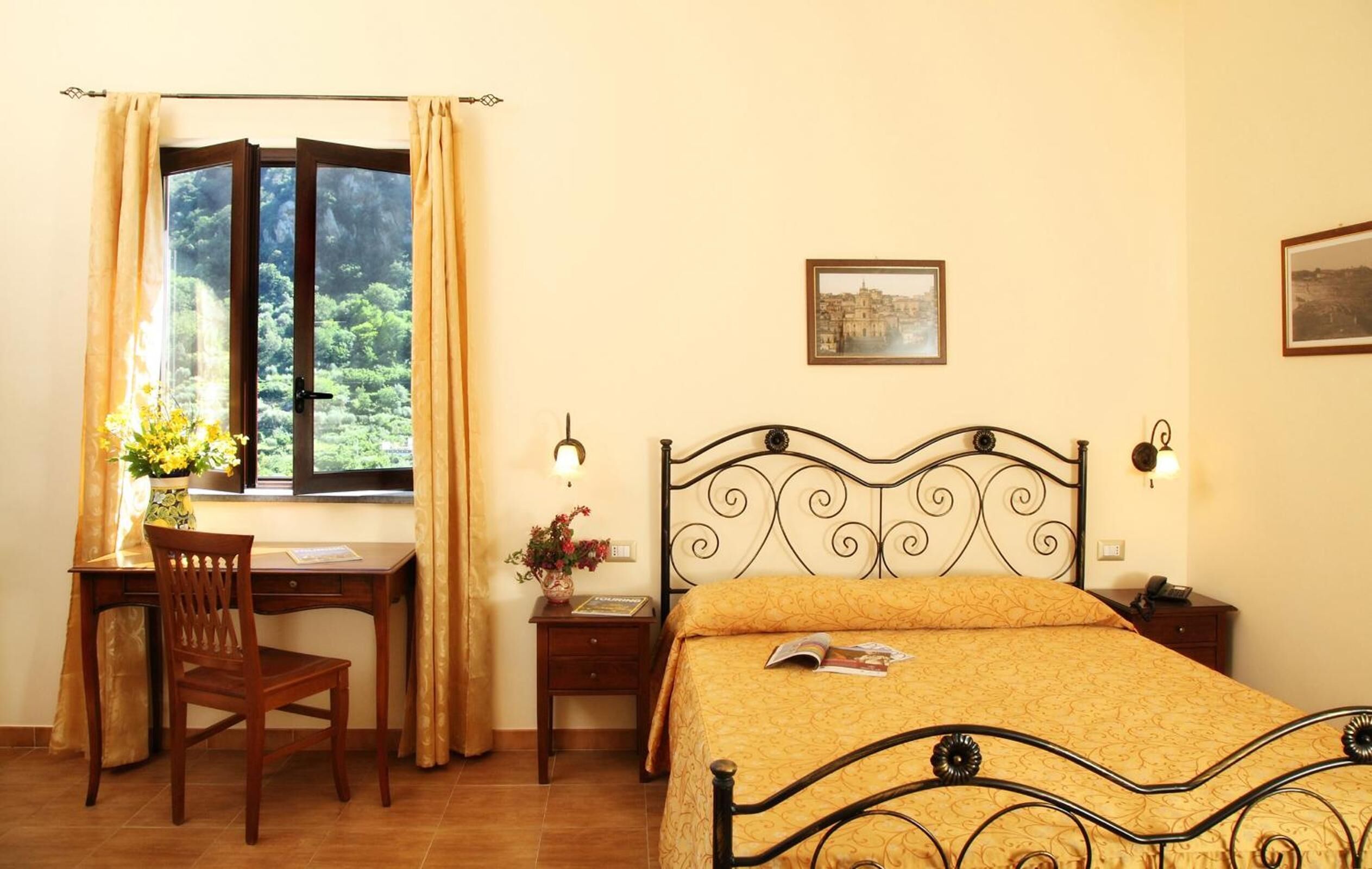 A bedroom with a bed, a desk, and a chair. There is a window with curtains, a framed picture on the wall, and a bedside table with a lamp.