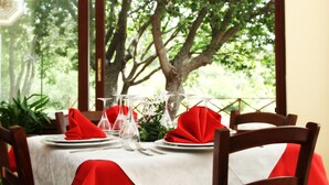 Mesa de comedor dispuesta con servilletas rojas, cristalería transparente y platos blancos, con vistas a árboles a través de una ventana.