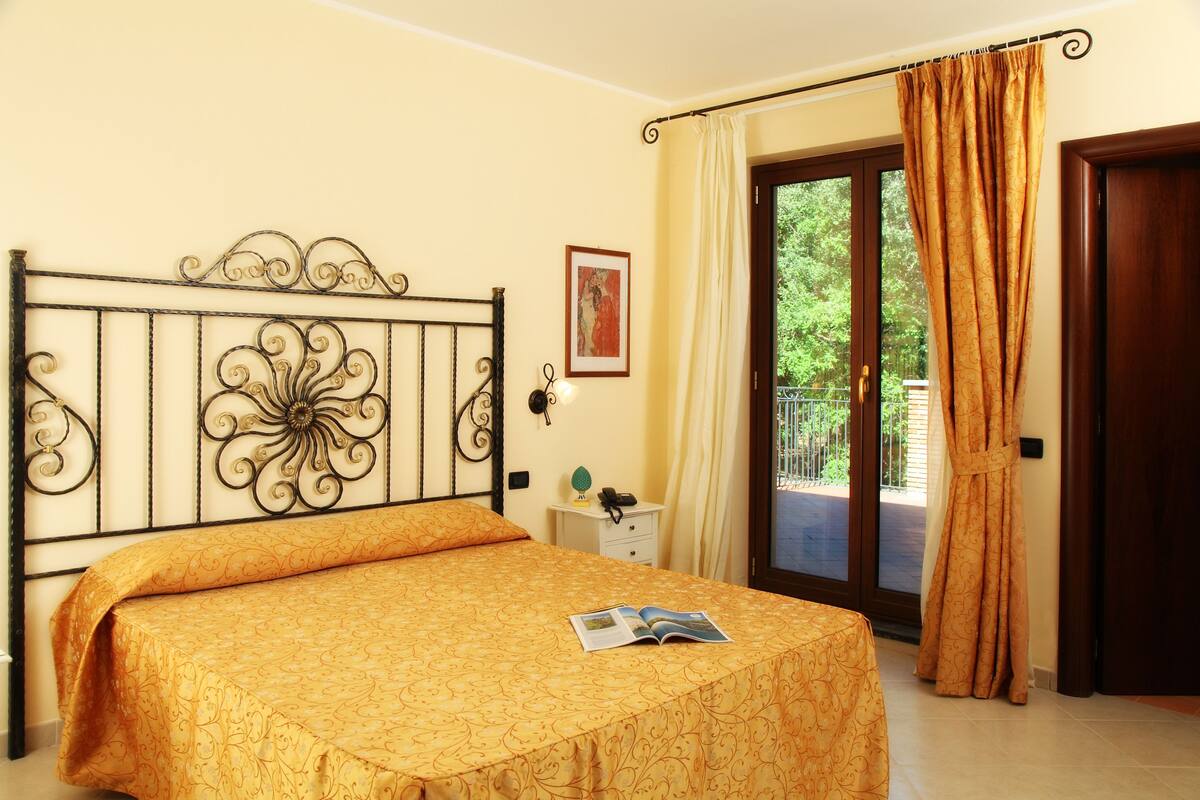 Quarto de hotel com cama, mesa de cabeceira com abajur, quadro na parede e janela com cortinas.