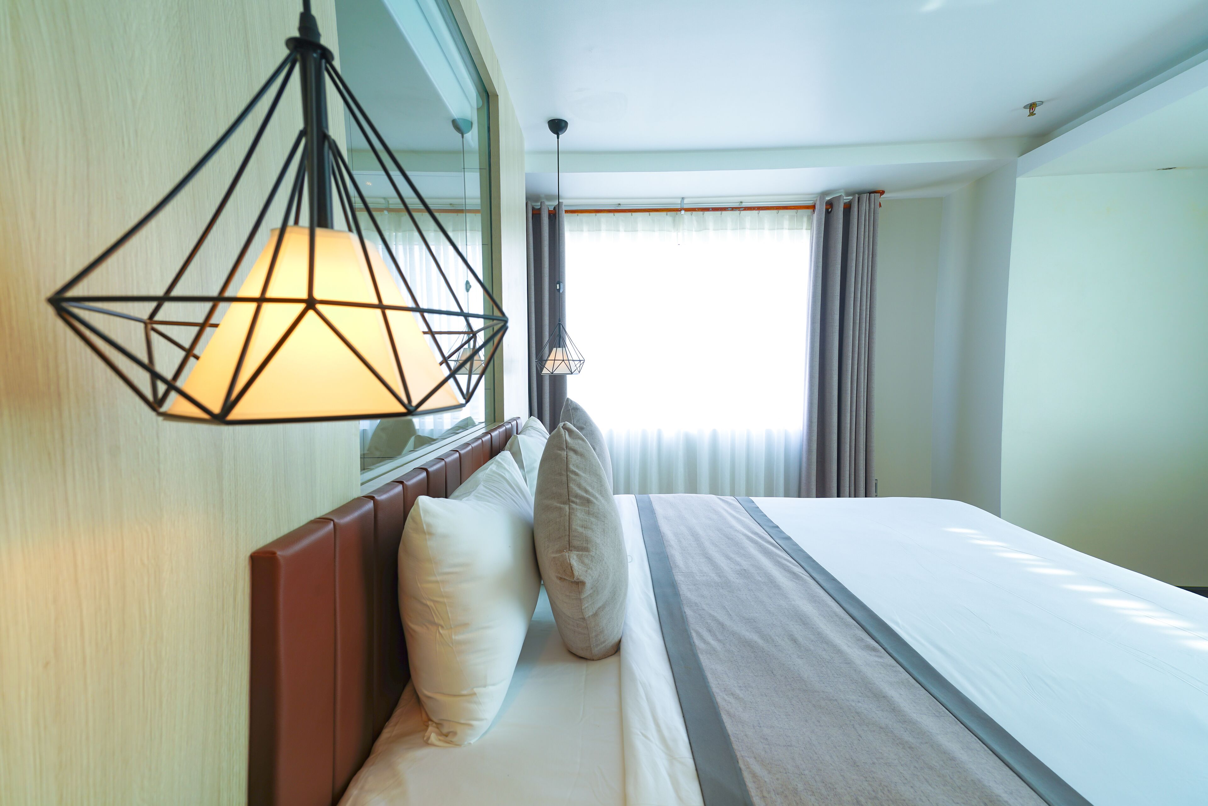 A hotel room with a bed, a hanging light fixture, a window with curtains, and a view of the outside.