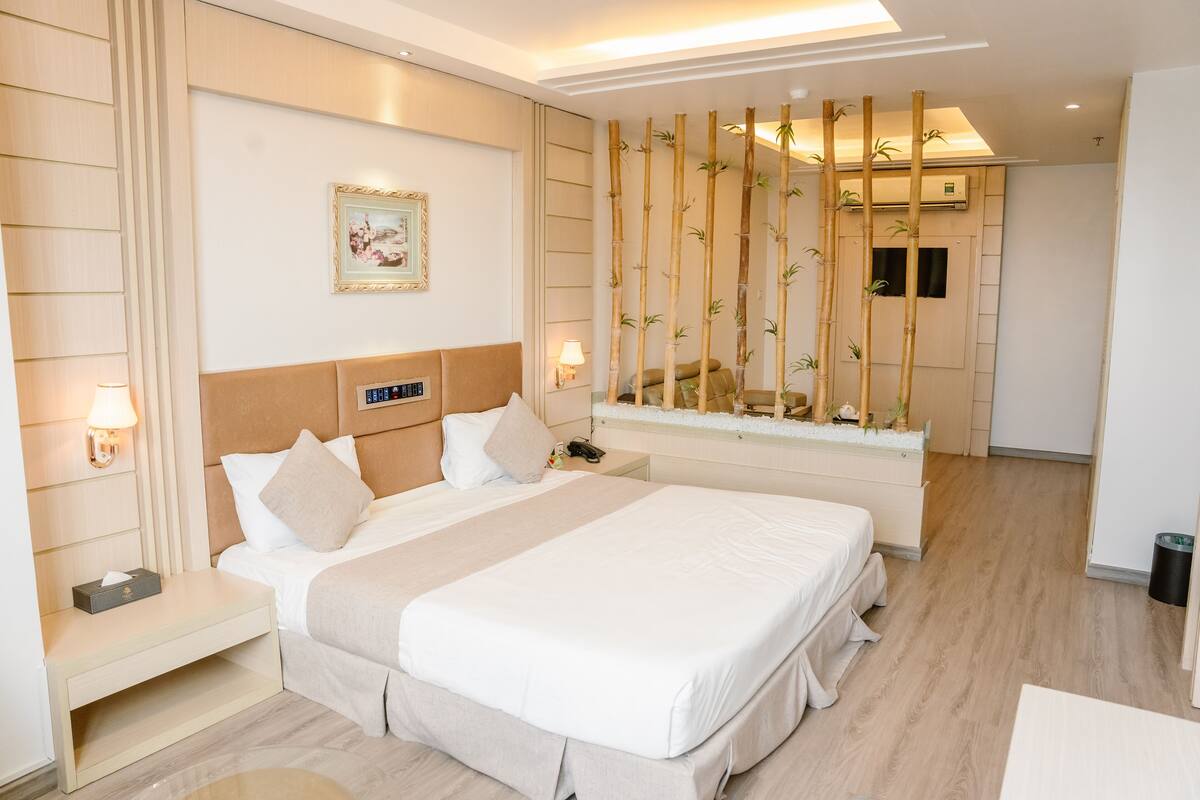 Une chambre d’hôtel moderne dotée d’un grand lit, de tables de chevet, d’un téléviseur fixé au mur et d’un paravent en bambou.