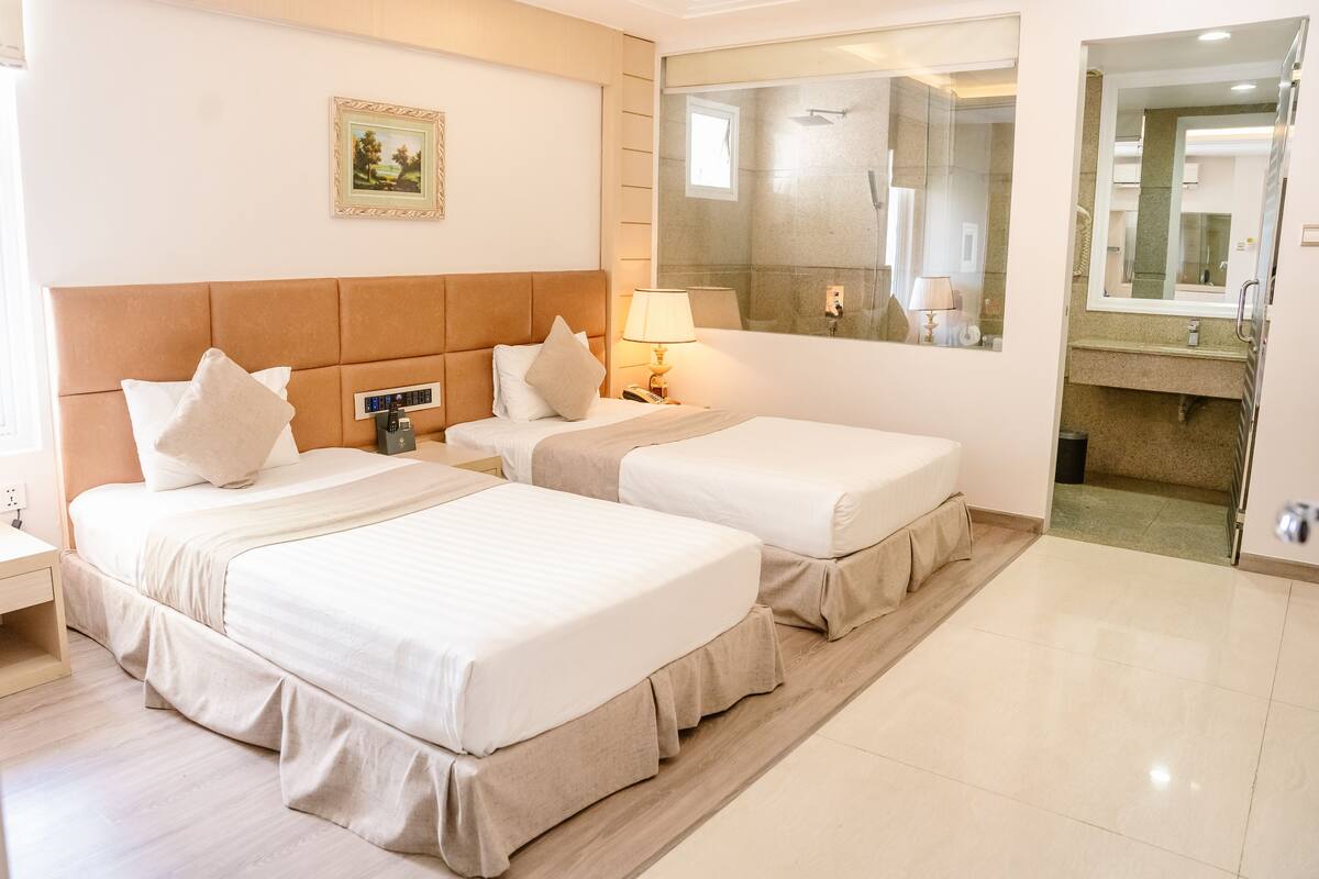 Une chambre d’hôtel avec deux lits, une table de chevet, une lampe, un grand miroir et une salle de bain visible à travers une porte ouverte.