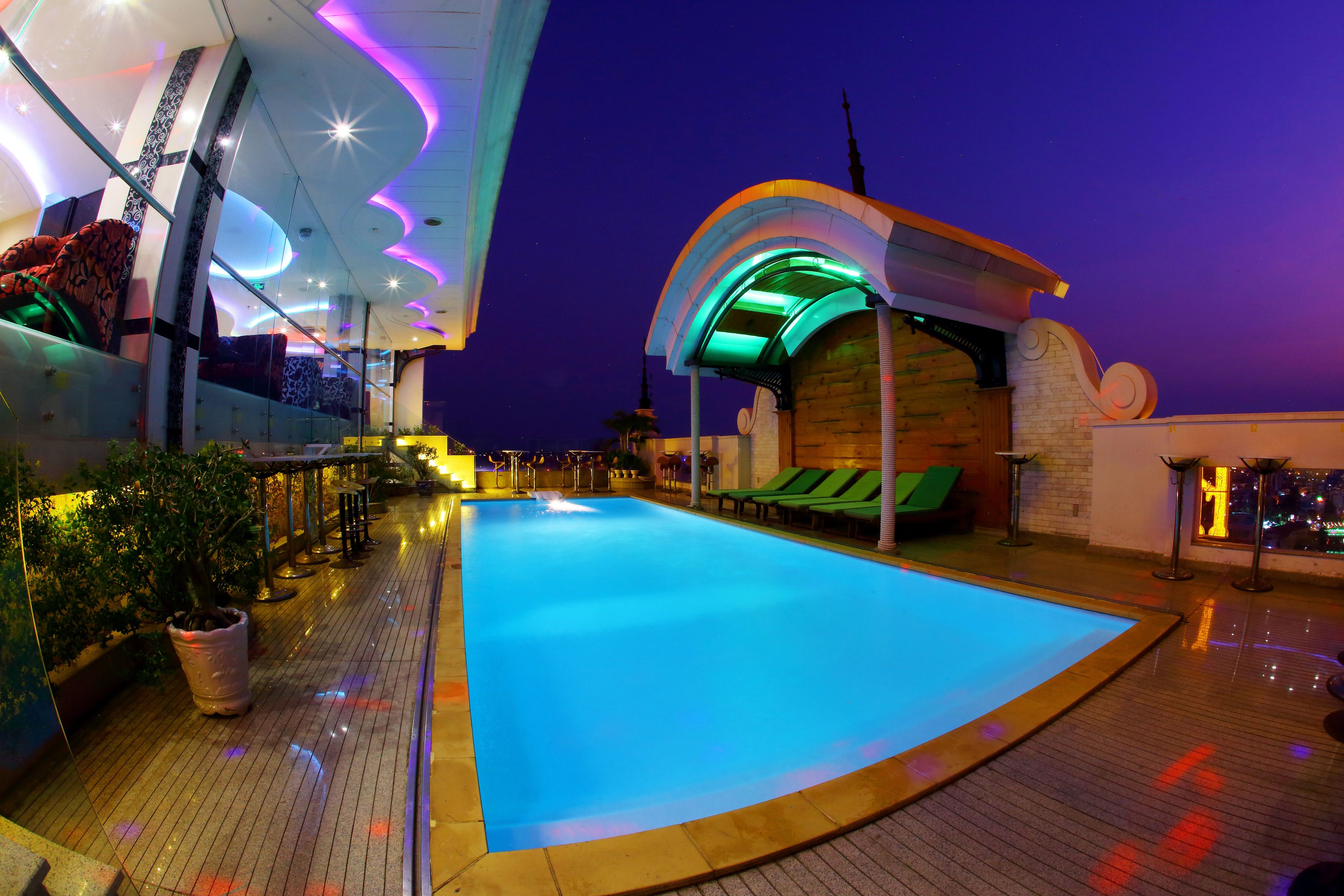Un área de piscina con piscina azul, sillas de descanso y una zona de estar cubierta con sillas verdes.
