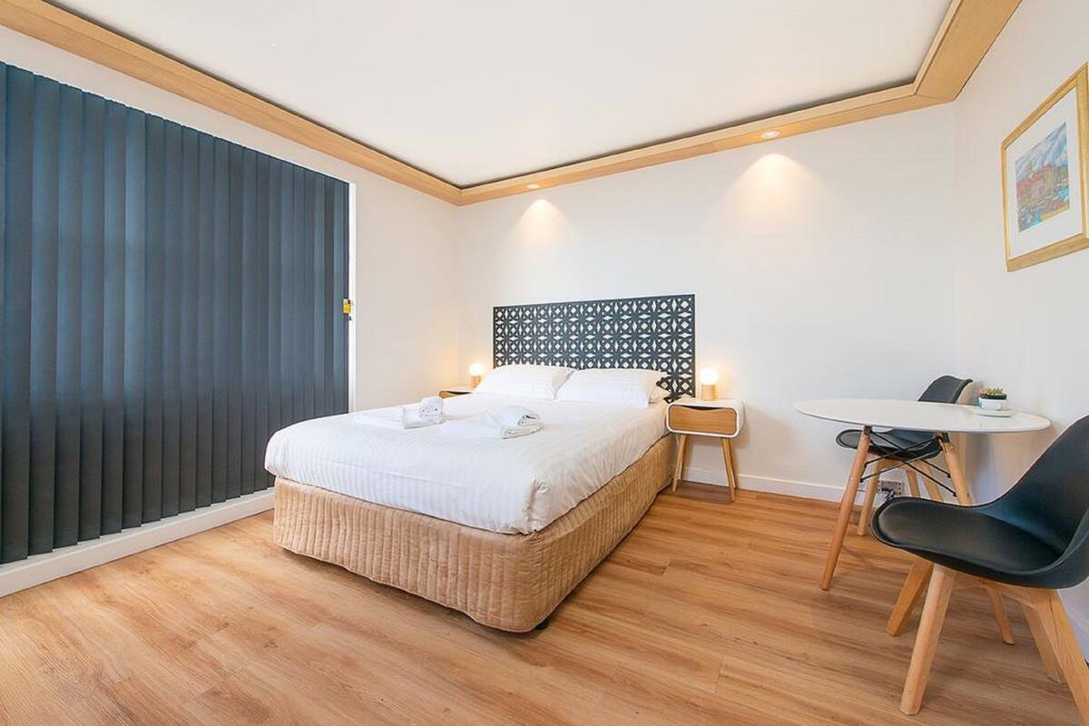A modern hotel room with a bed, bedside table, chair, and a small table with a lamp.