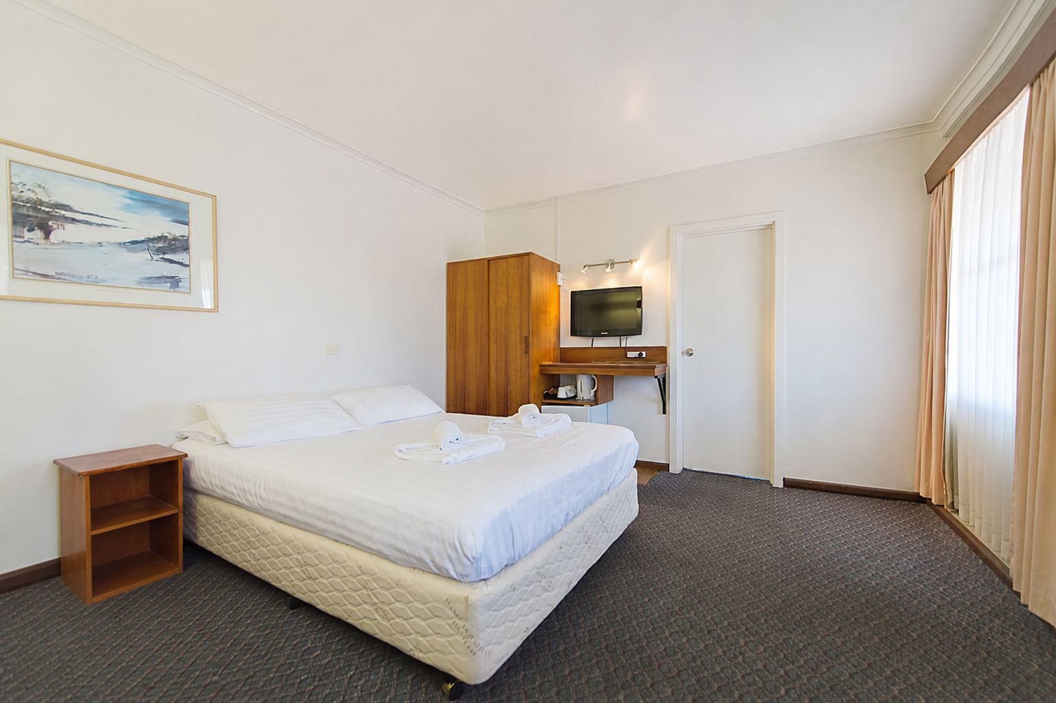 A hotel room with a bed, a television, a wooden wardrobe, a small table, and a painting on the wall.