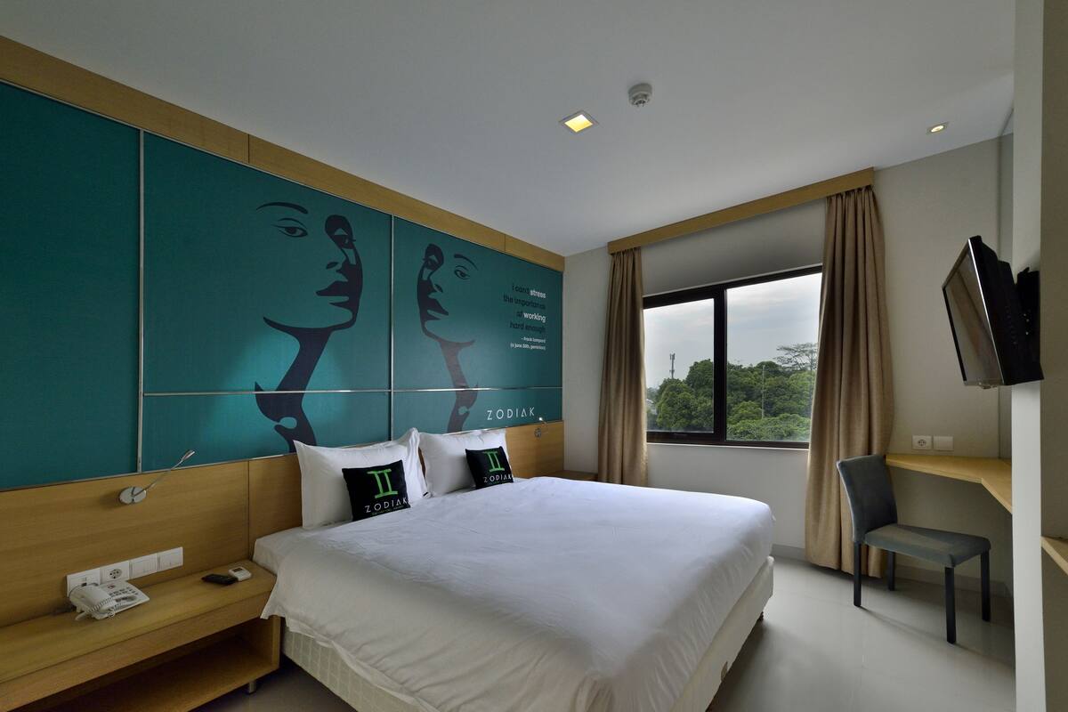 A hotel room with a large bed, a desk with a chair, a TV, and a window with curtains.