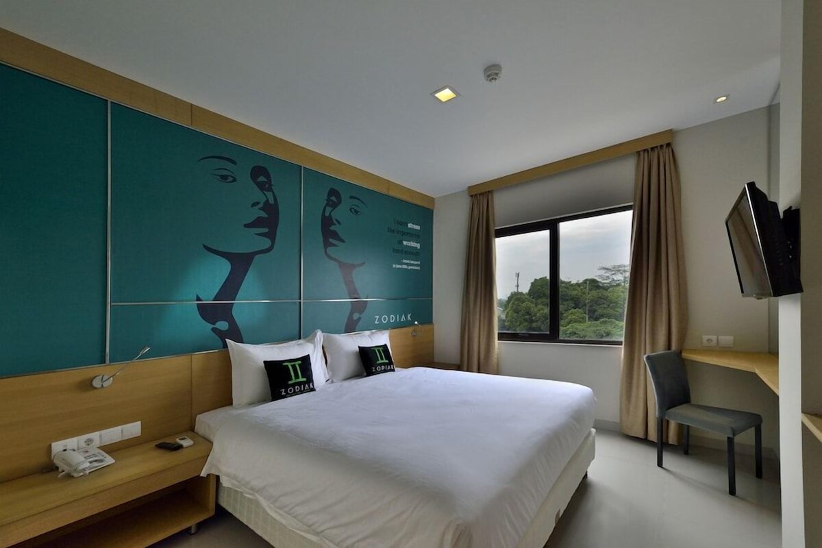 A hotel room with a large bed, a desk with a telephone, a chair, a TV mounted on the wall, and a window with a view of trees.