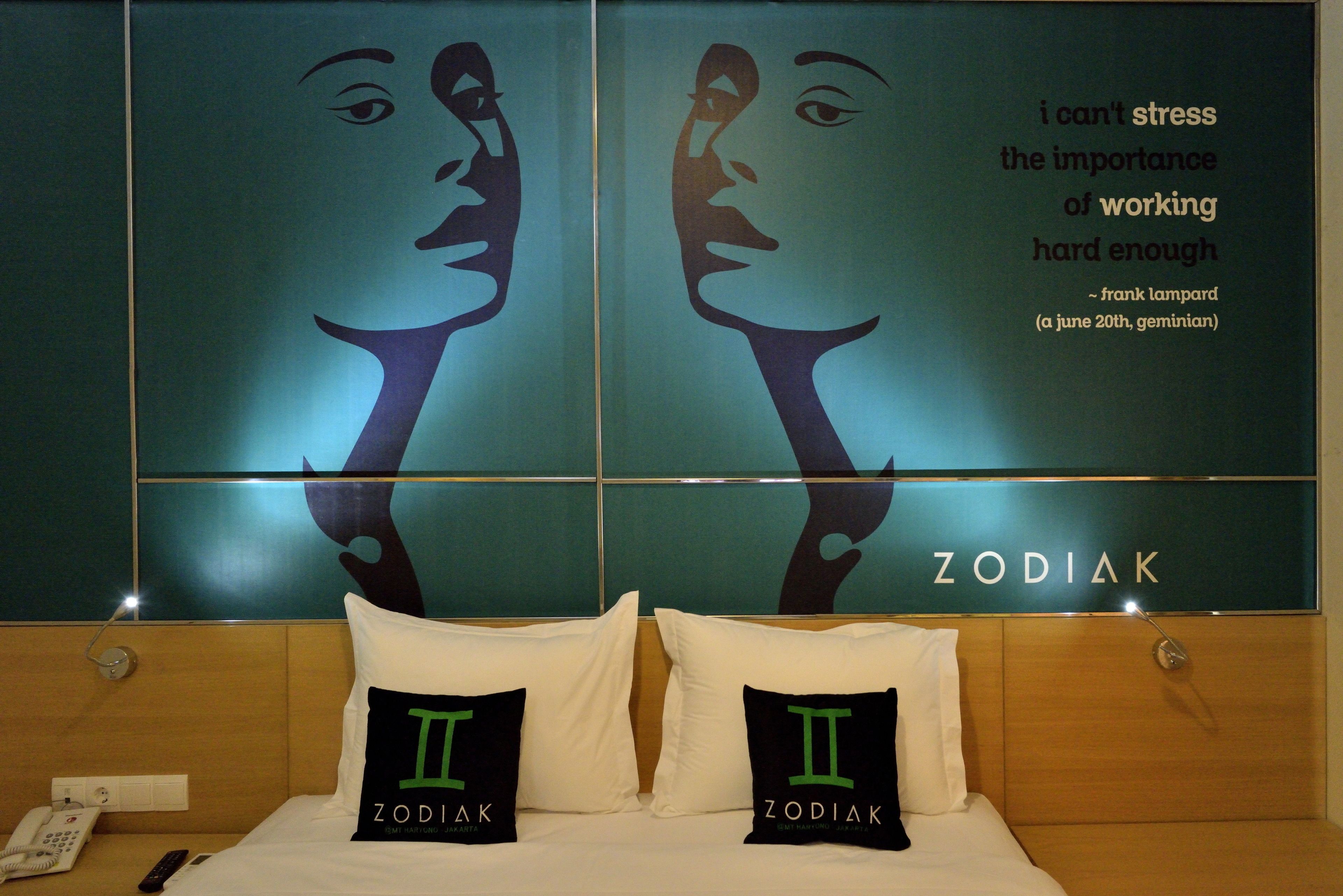 A hotel room with a bed featuring two pillows with the Zodiac sign II, a poster with a quote, and a sign above the bed that reads "ZODIAK".