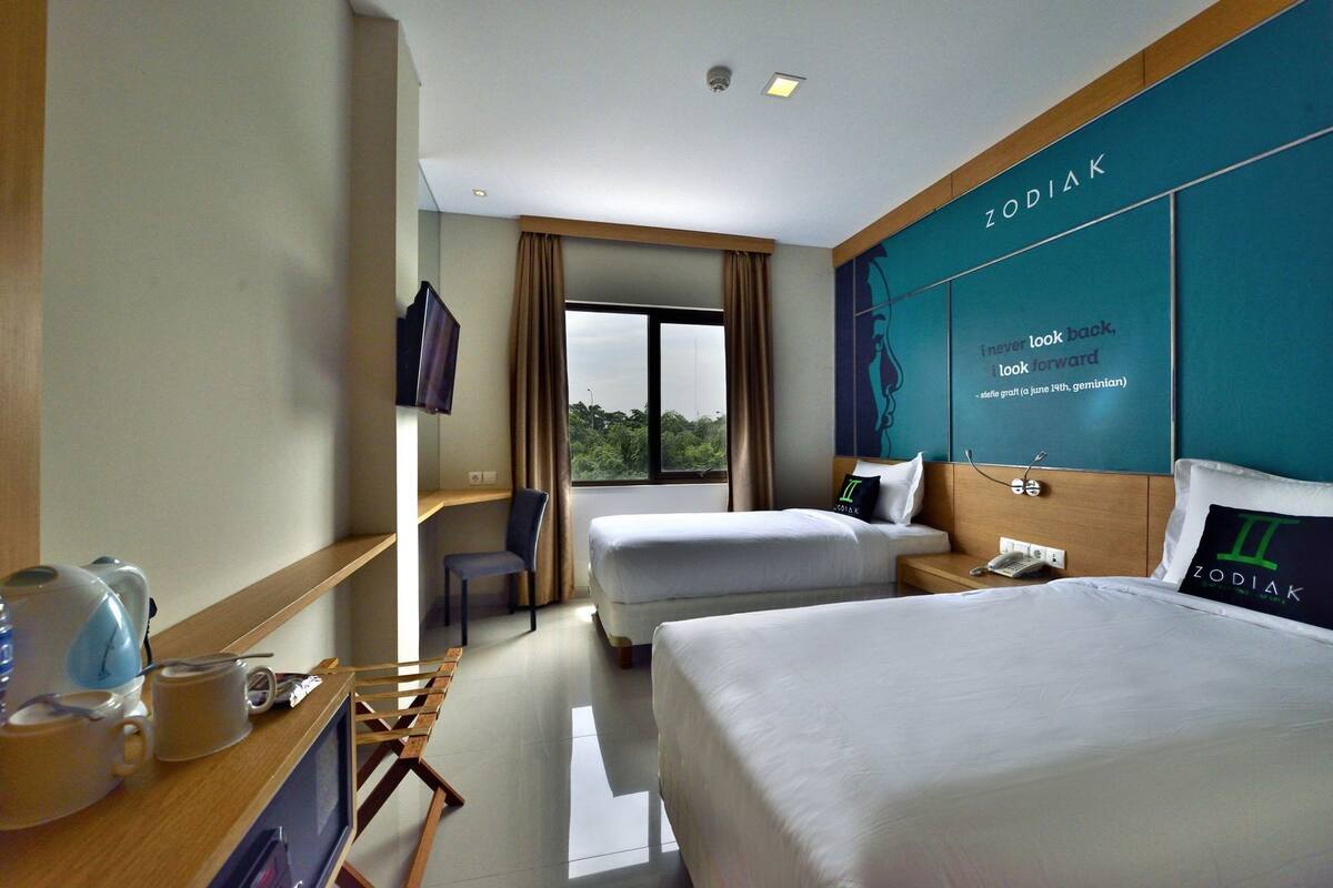A hotel room with two beds, a desk with a chair, a TV, and a large window.