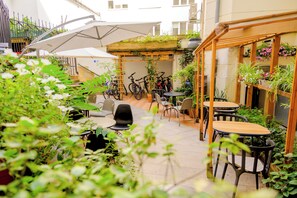 An outdoor patio area with multiple tables and chairs, surrounded by potted plants and a wooden pergola.