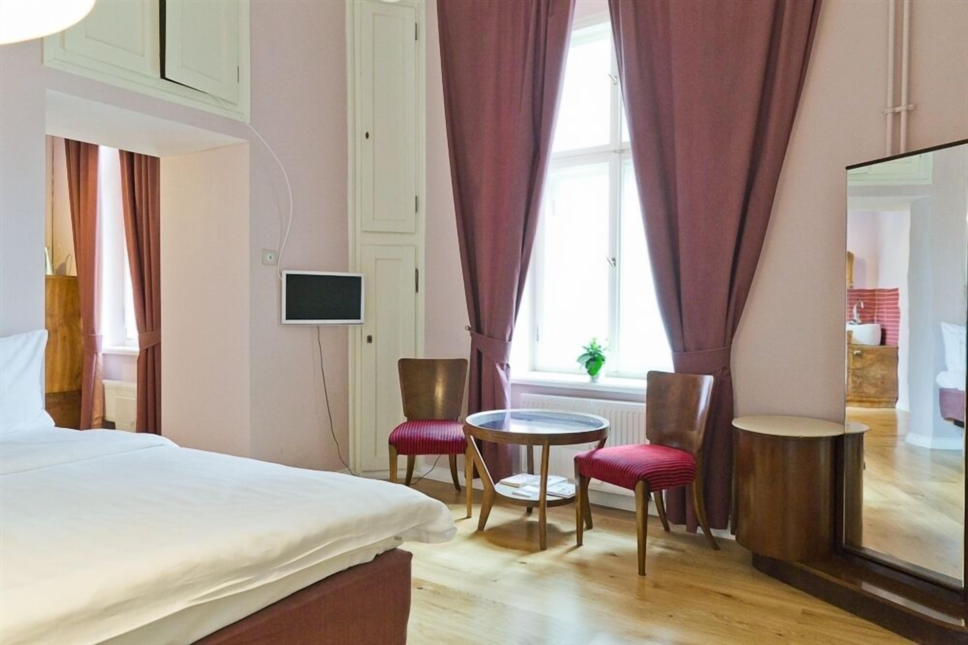 Een hotelkamer met een bed, twee stoelen, een klein rond tafeltje, een televisie en een raam met gordijnen.