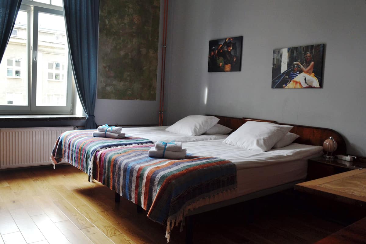 A hotel room with a large bed, a wooden floor, a window with curtains, and two paintings on the wall.