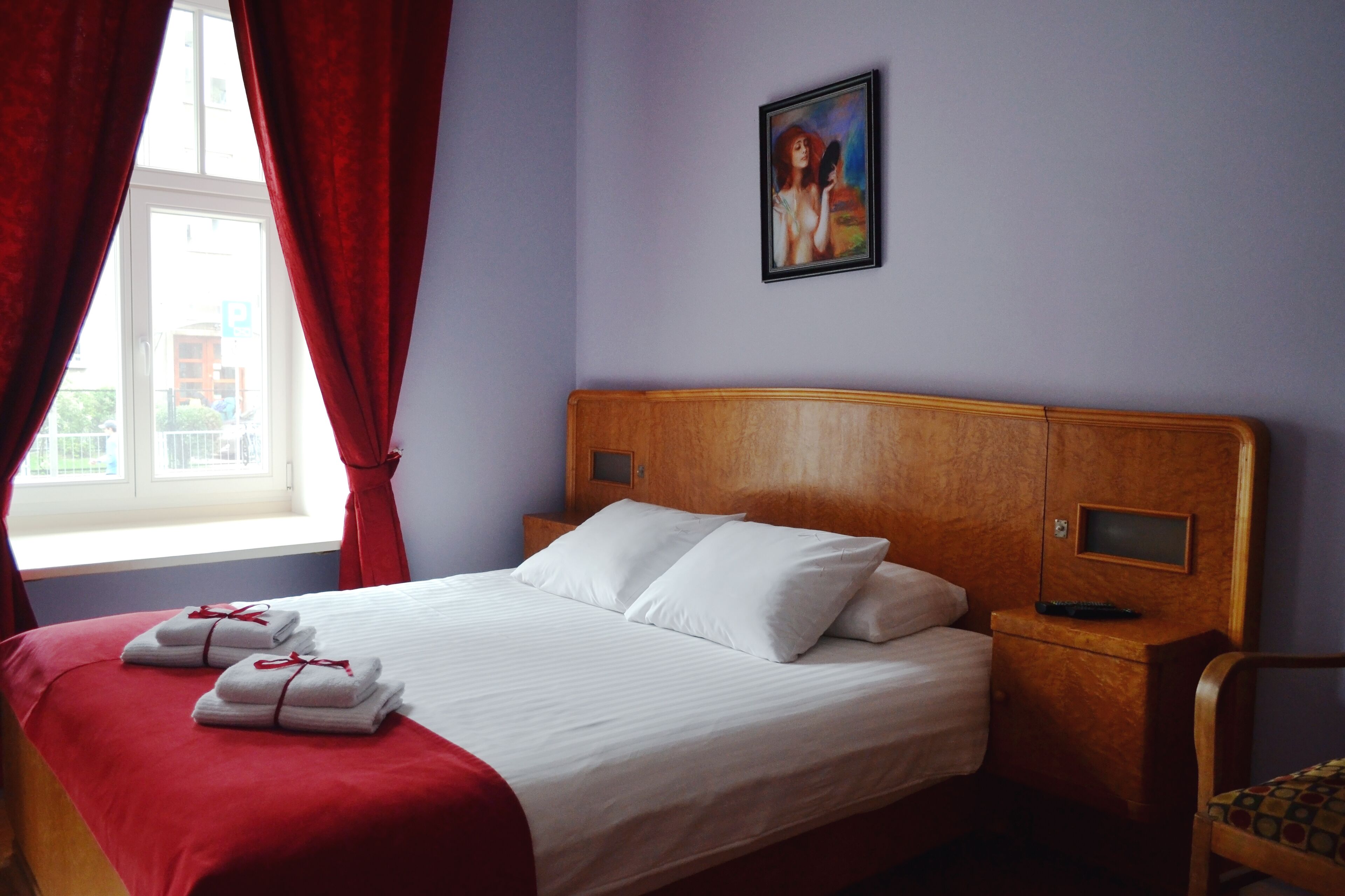 Une chambre d’hôtel avec une tête de lit en bois, du linge de lit blanc, des rideaux rouges, un tableau au mur et une fenêtre donnant sur des immeubles.