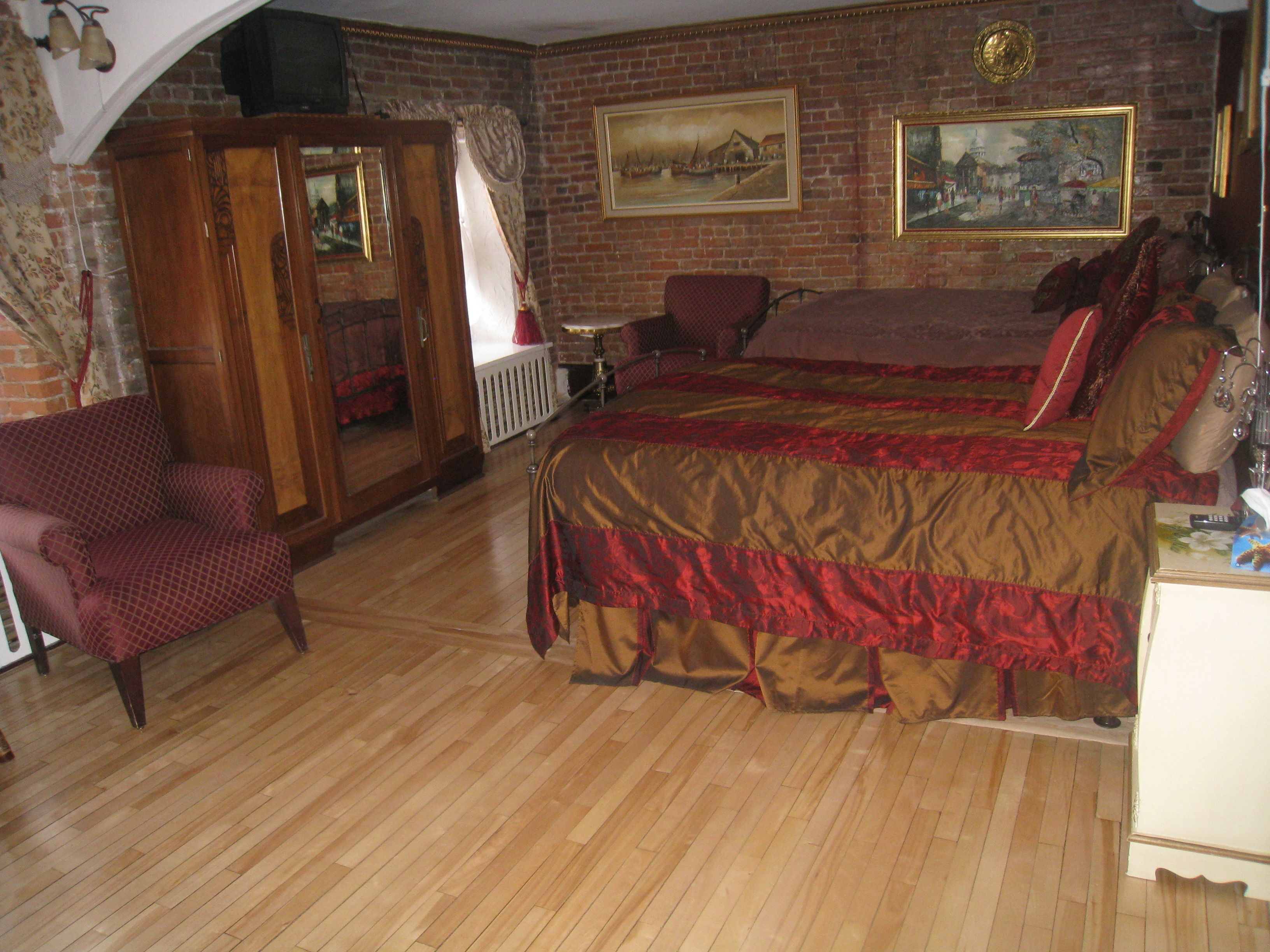 A bedroom with a wooden floor, a large bed with red and gold bedding, a wooden wardrobe, a red armchair, and two paintings on the brick wall.
