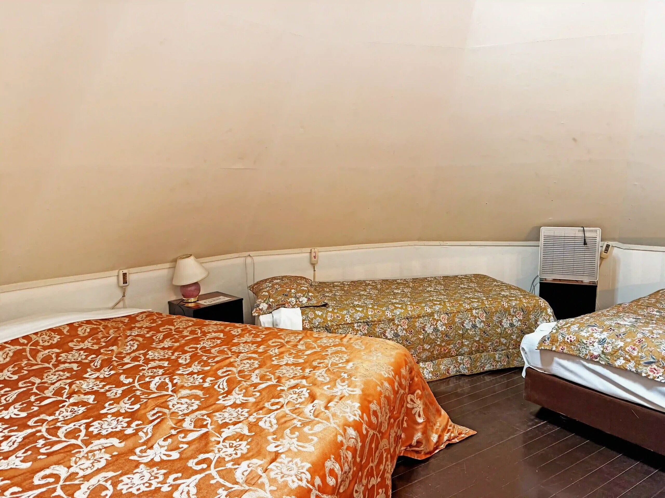 A room with three beds, each with a different patterned bedspread. There is a bedside table with a lamp and a power outlet. An air conditioning unit is mounted on the wall.