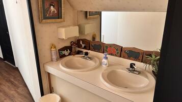 Une salle de bain avec deux lavabos, un miroir, un tableau encadré au mur et une petite poubelle.