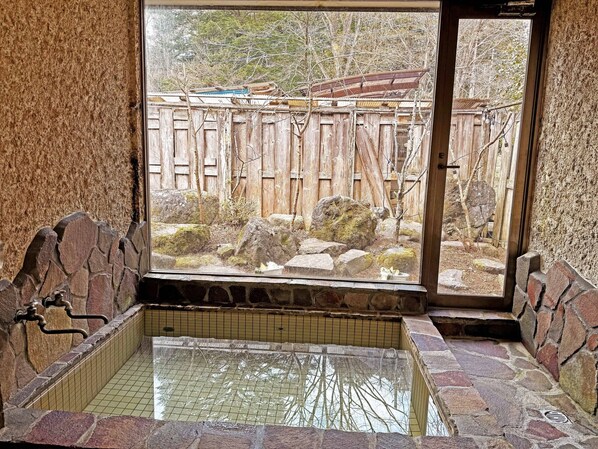 Une baignoire en pierre, un sol carrelé, une clÎture en bois et une vue sur un jardin.