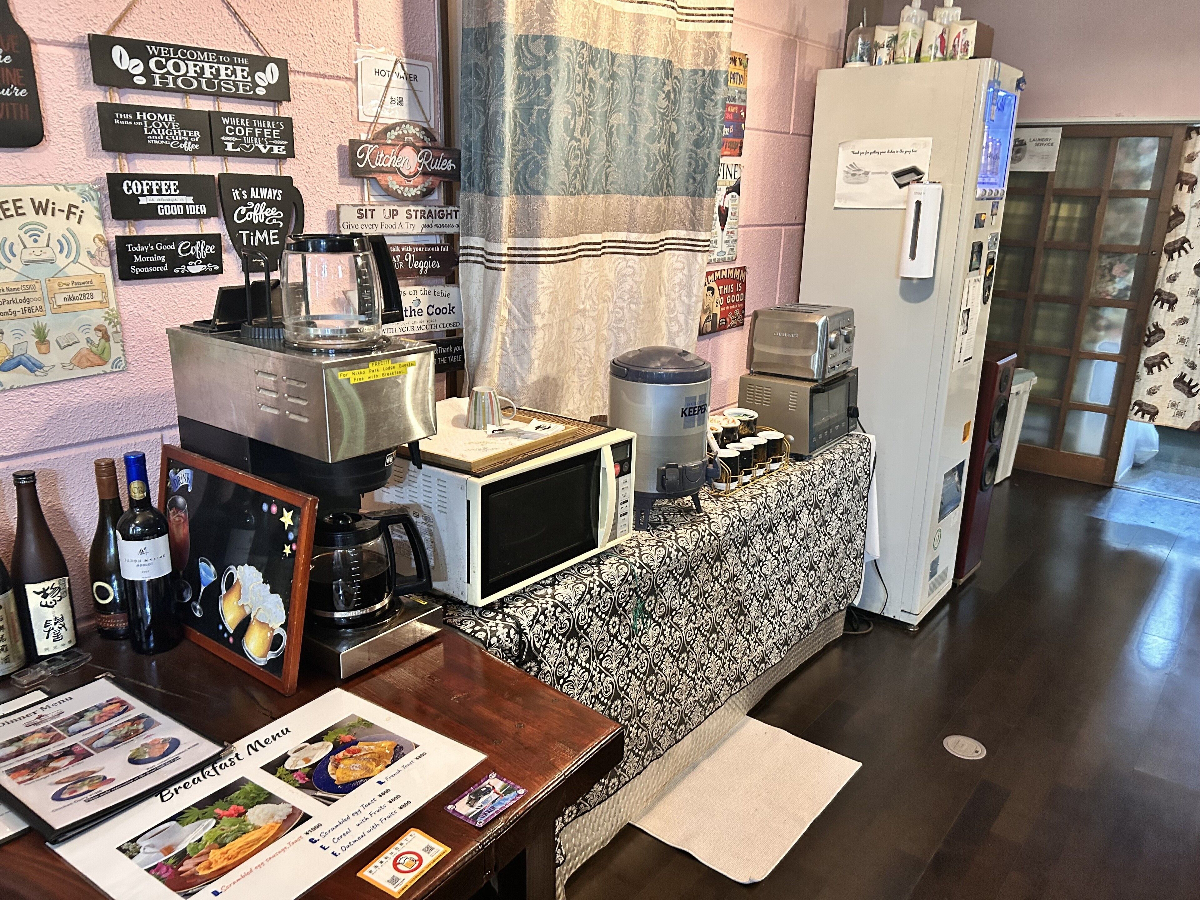 Ein Küchenbereich mit Kaffeemaschine, Kühlschrank, Mikrowelle und verschiedenen Küchengeräten. An der Wand befinden sich Schilder und auf der Arbeitsplatte ein Menü.