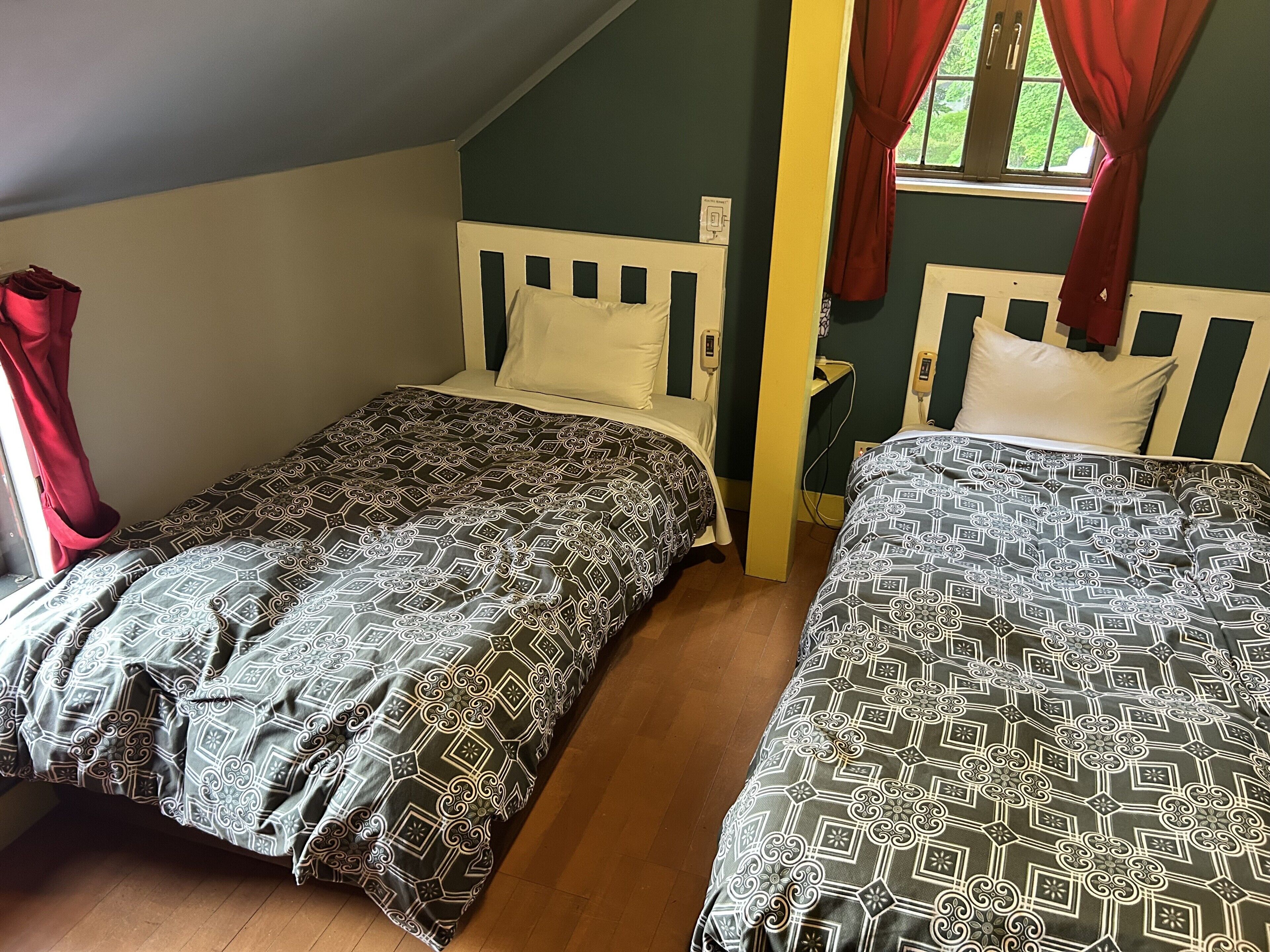 Two single beds with patterned bedding, a wooden floor, a window with red curtains, and a green wall.