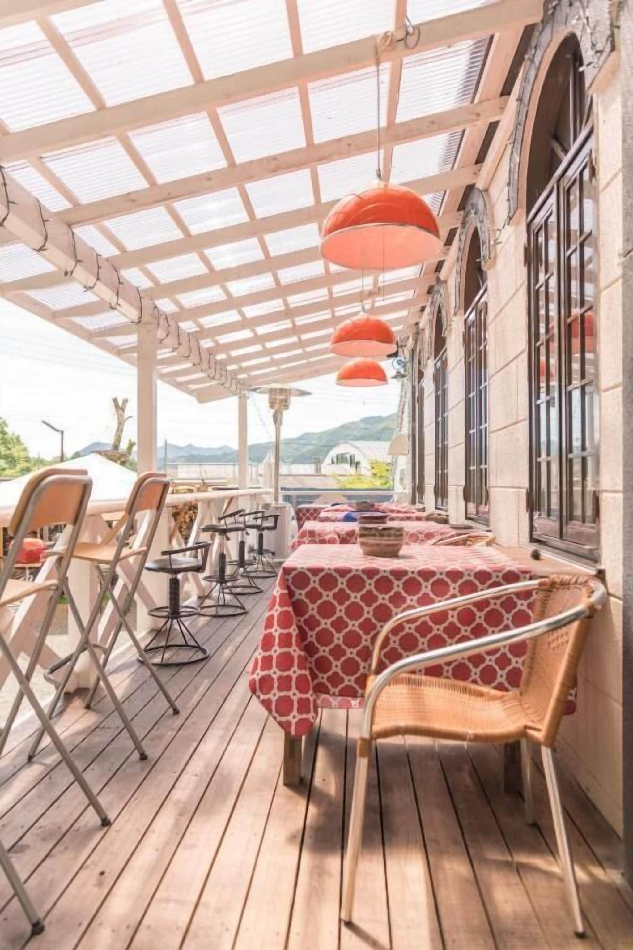 A covered outdoor dining area with tables and chairs, a pergola with orange lamps, and a view of the landscape.